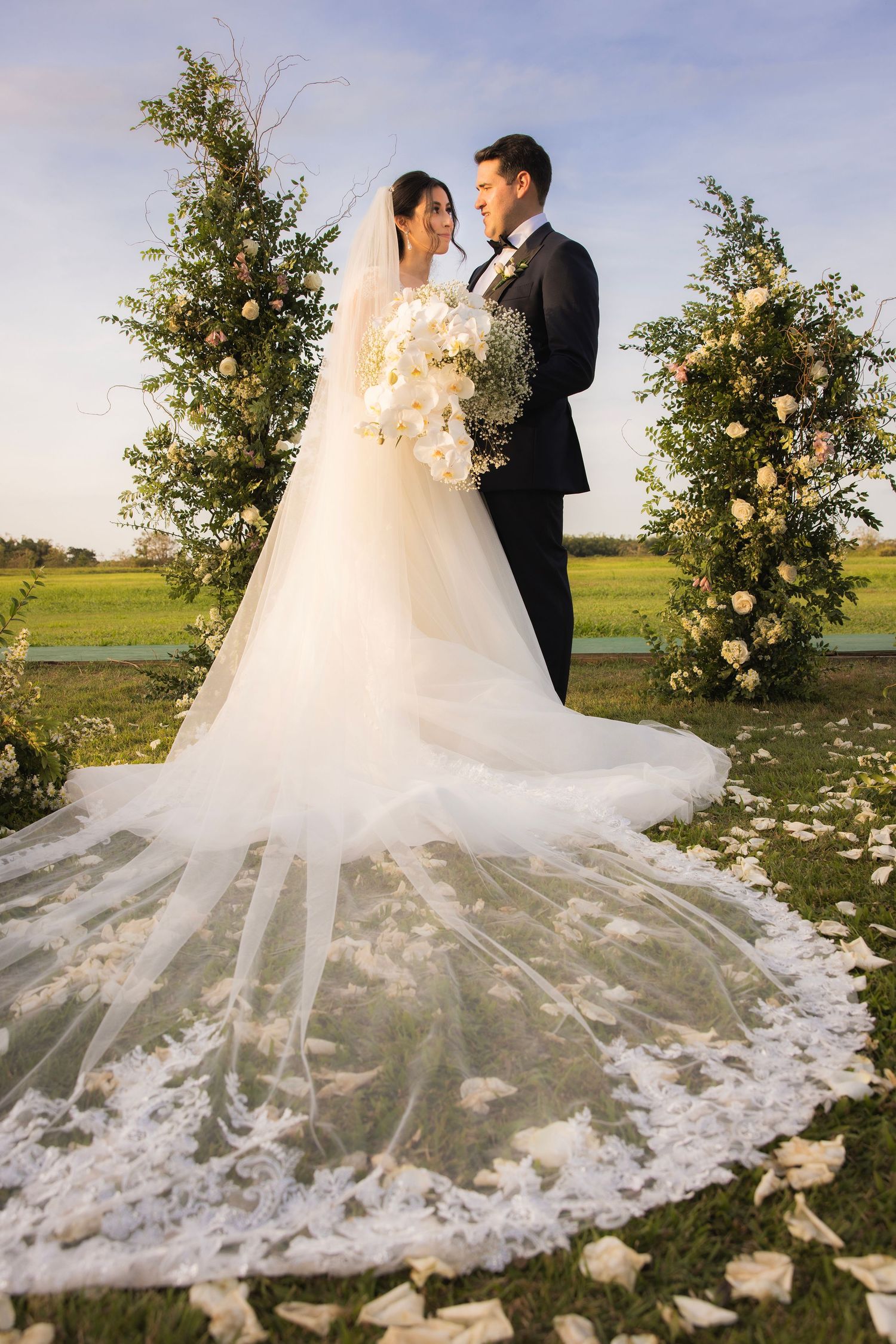 Beautiful wedding portrait with a dramatic cathedral veil spread out over flower petals in an outdoor setting.