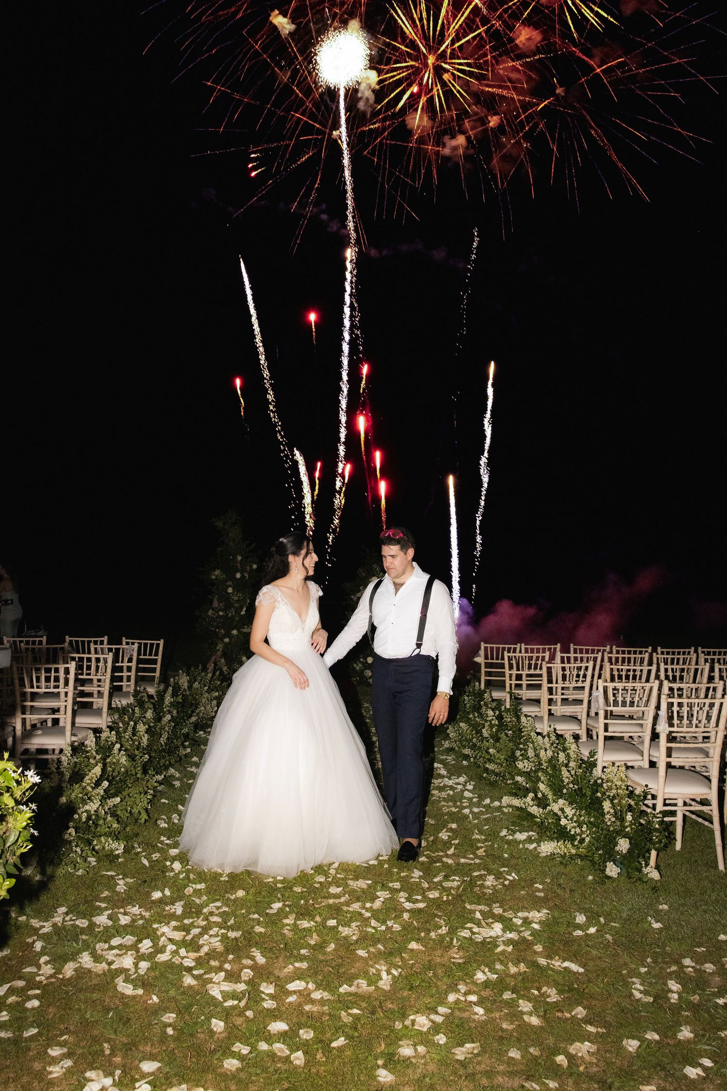 Couple enjoying a romantic moment under bursting fireworks during an outdoor evening wedding celebration with white chairs.