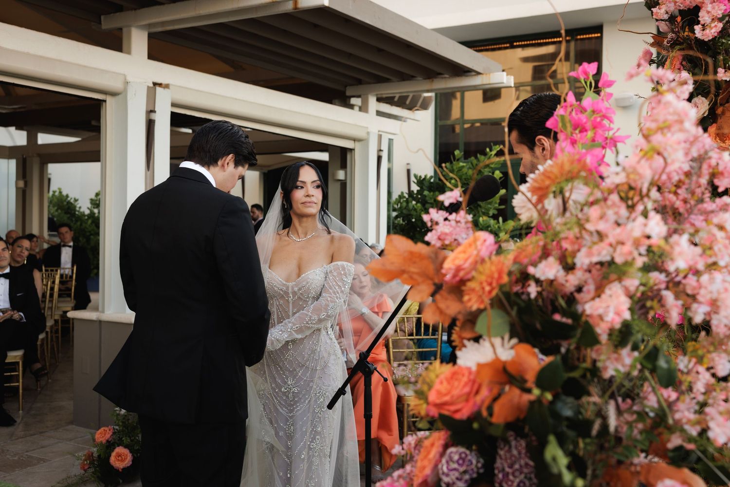 A wedding couple exchanges vows on a terrace decorated with vibrant pink and coral floral arrangements.
