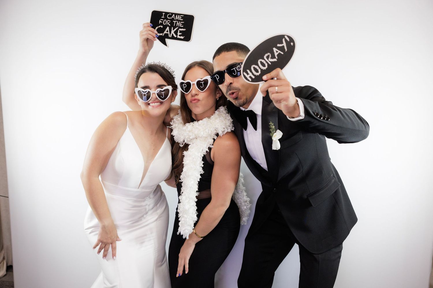 Four people in formal attire pose playfully with photo booth props against a white backdrop at a celebration event.