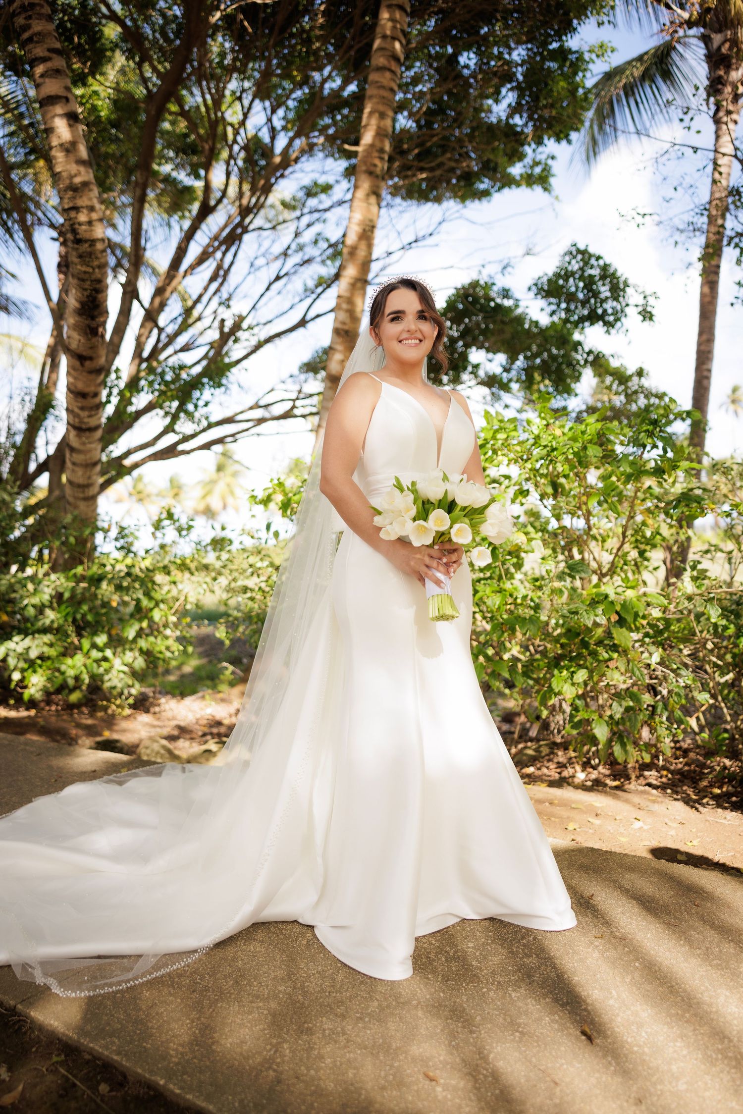 A bride in a flowing white wedding dress poses gracefully among tropical palm trees and lush landscaping.