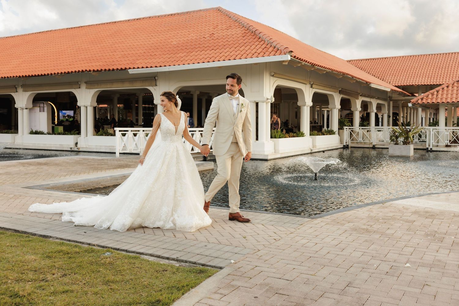 A couple in wedding attire strolls along a paved walkway beside a Spanish-style building with arched columns and fountain.