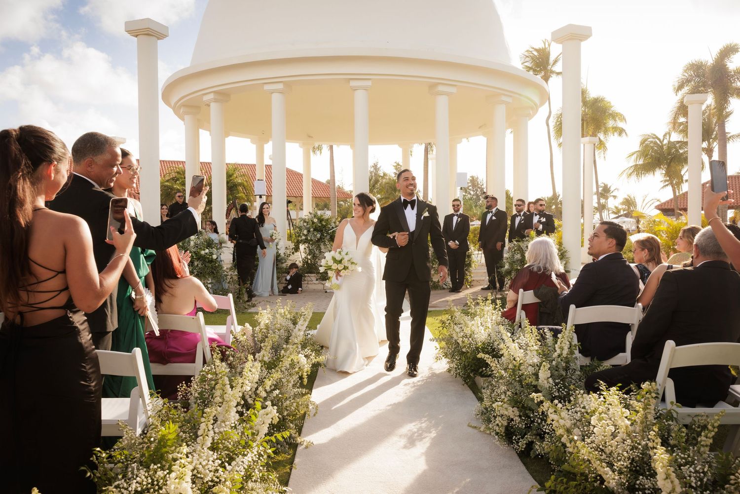 Wedding ceremony takes place under an elegant white domed gazebo with palm trees and sunlight in the background.