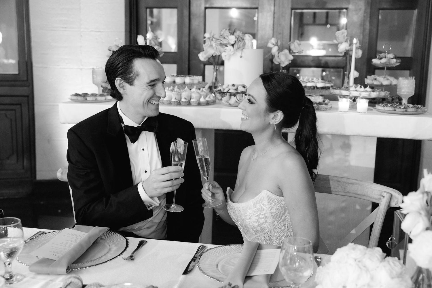 A black and white photo of a bride and groom sharing a toast at their wedding reception table.