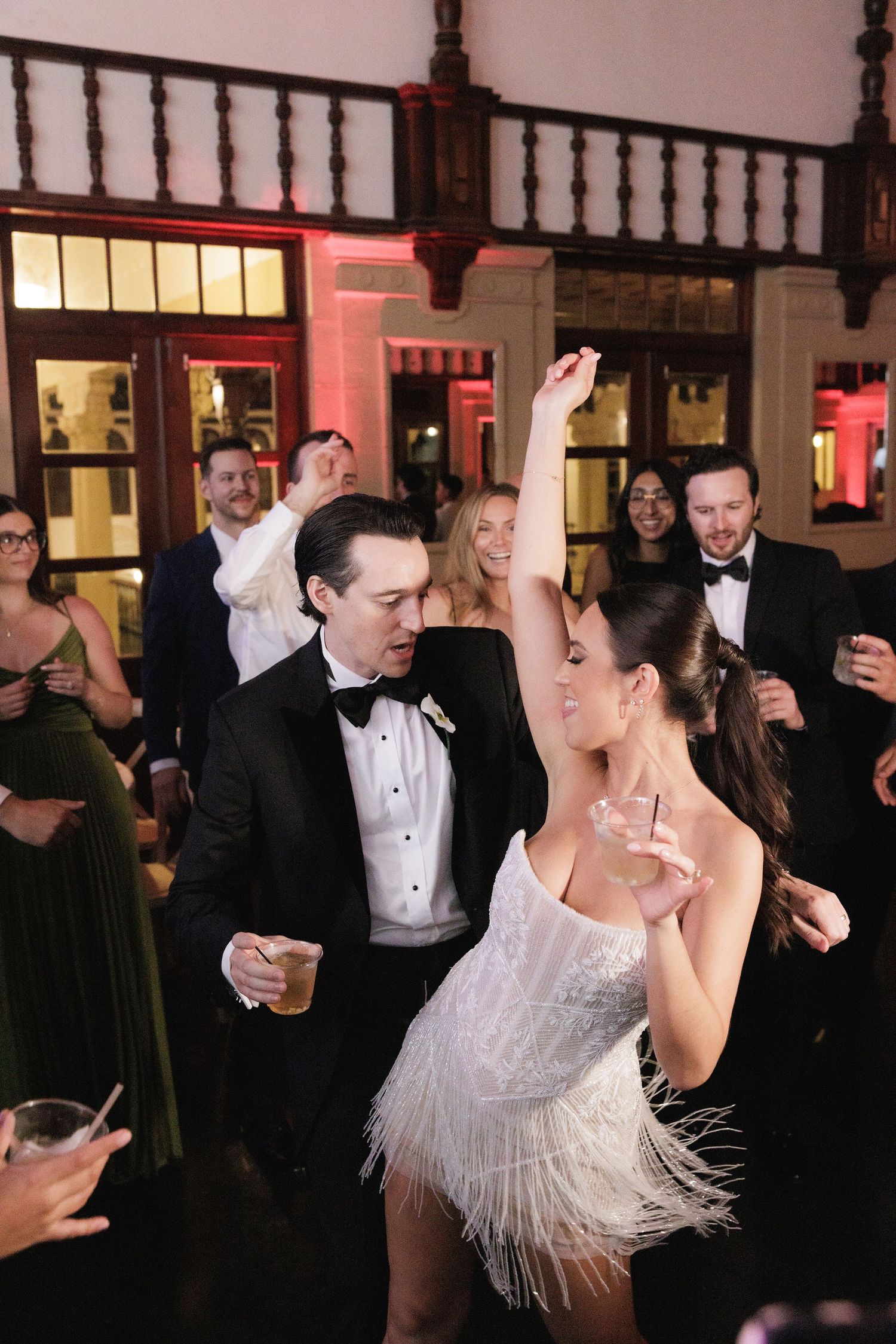 Wedding guests dance enthusiastically on a dance floor during an elegant reception with festive lighting and decor.