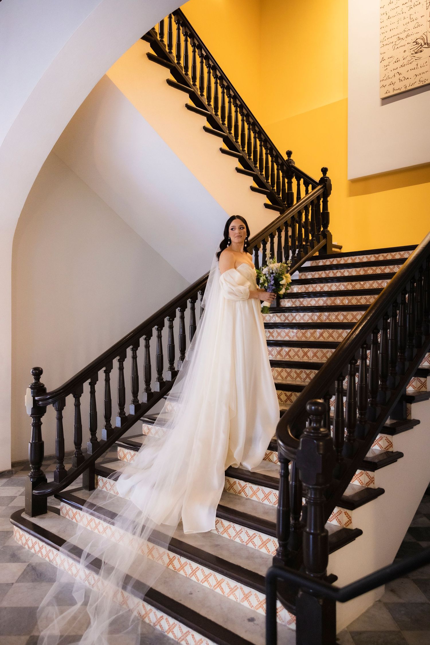 Wedding dress train cascades down a dark wooden staircase with warm lighting.