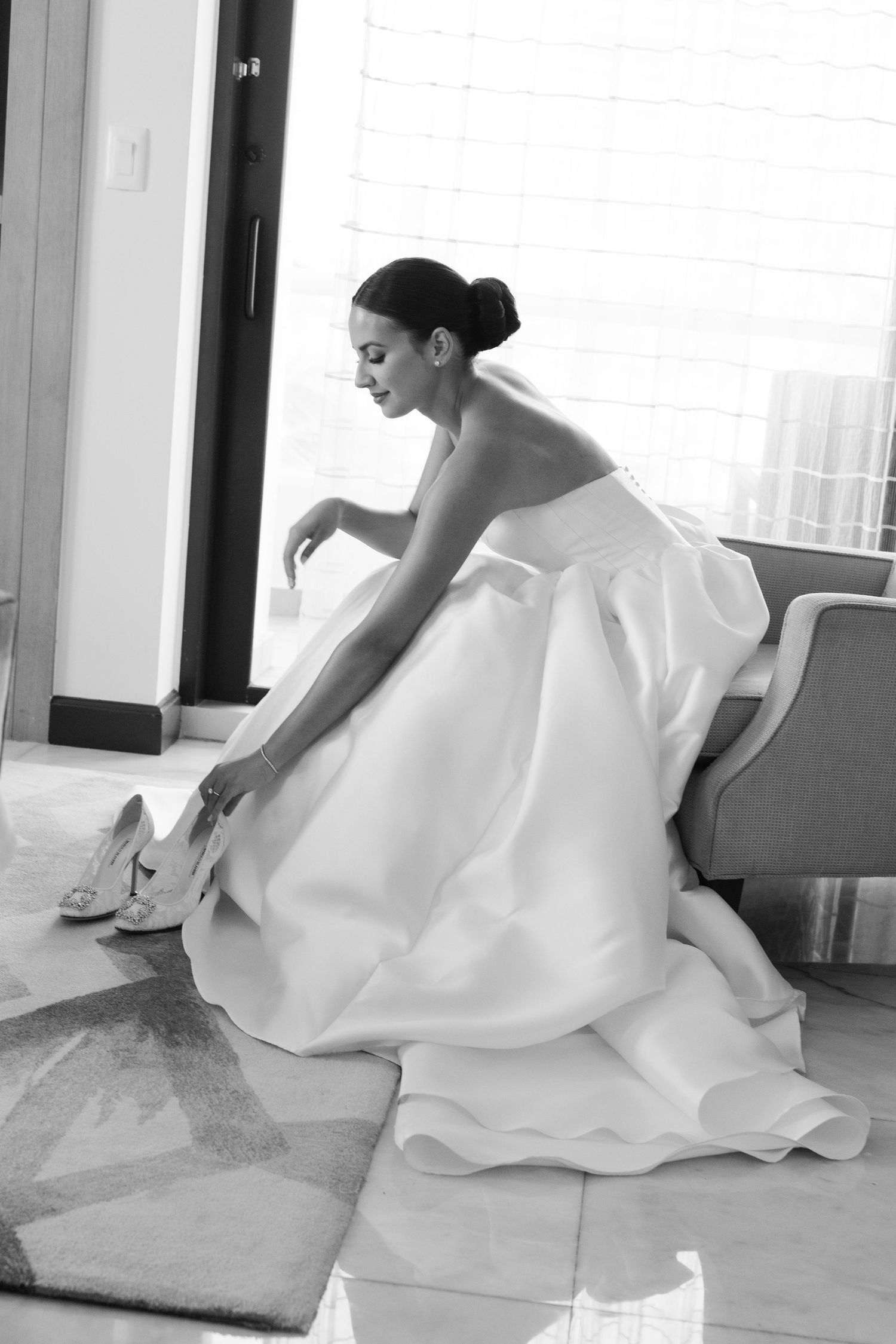 Bride in flowing white dress adjusting shoes in sunlit room.