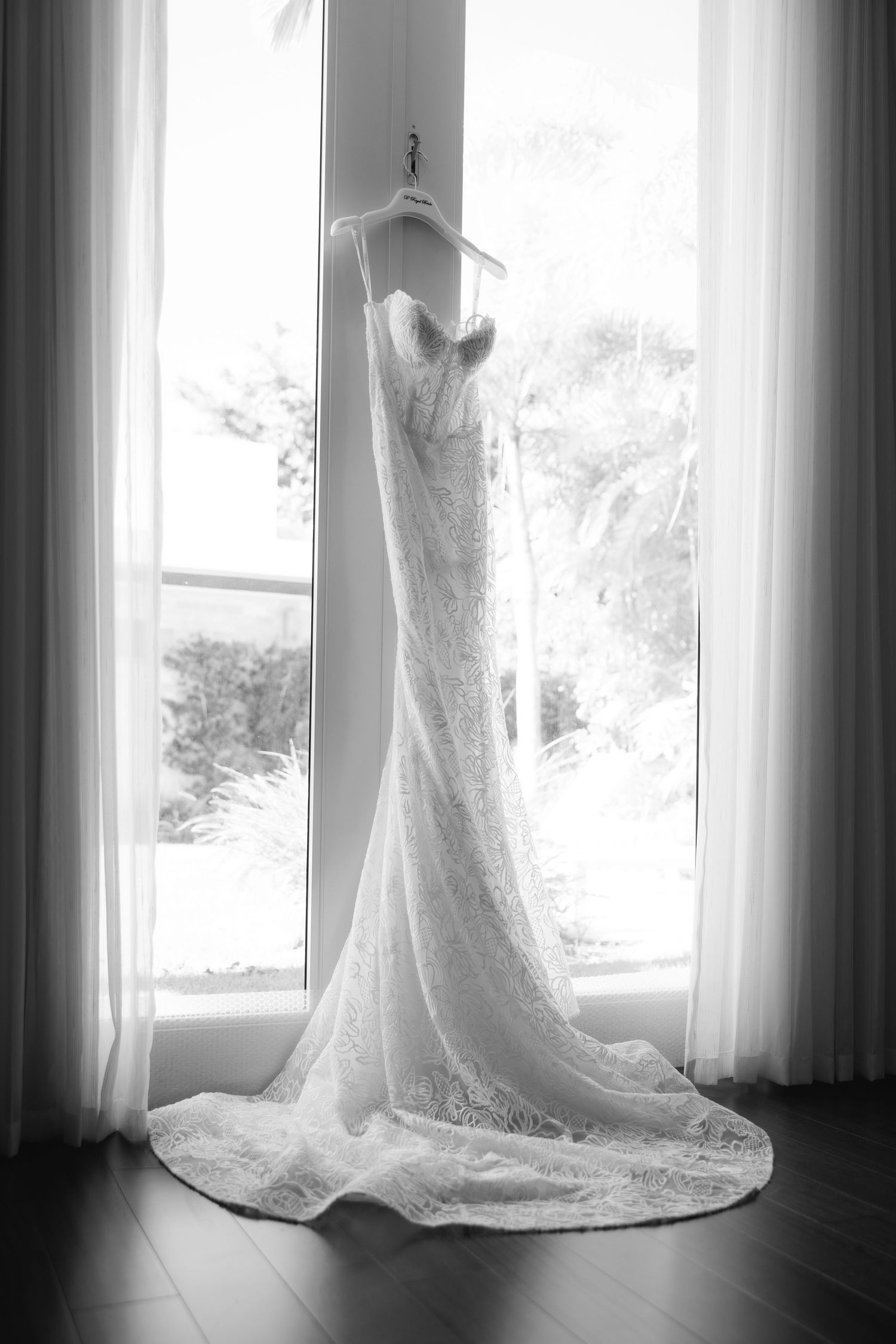 Elegant white lace wedding dress with long train hanging by window in dramatic black and white lighting.