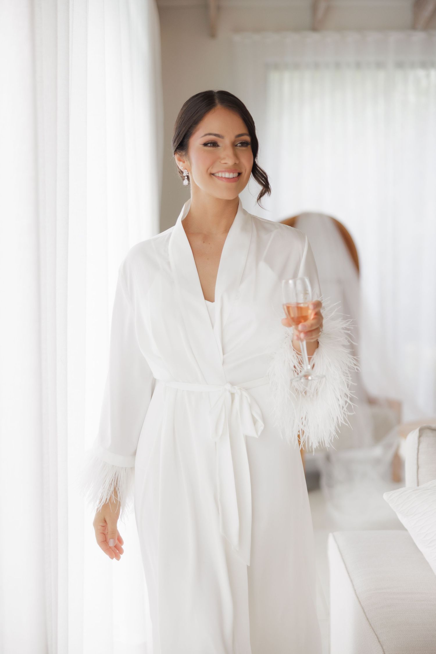 A bride in an elegant white robe poses with a champagne glass in a bright, airy room.