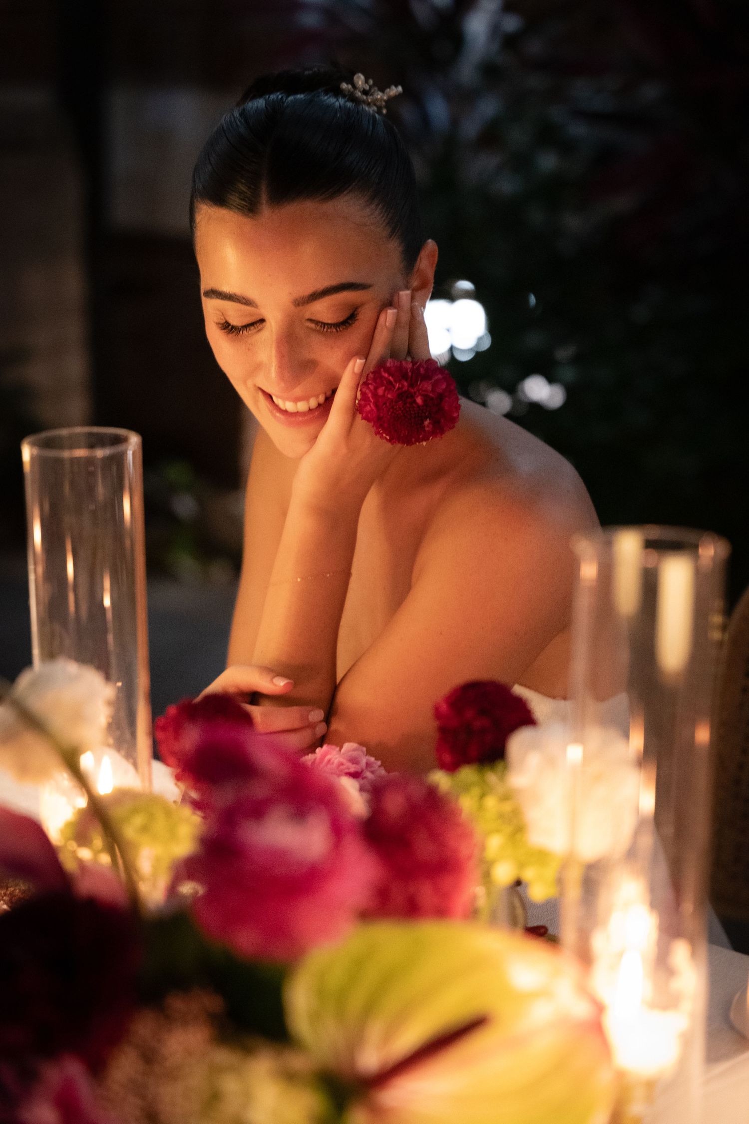 Smiling bride with floral earring leans on hand at candlelit table surrounded by pink and white flowers.