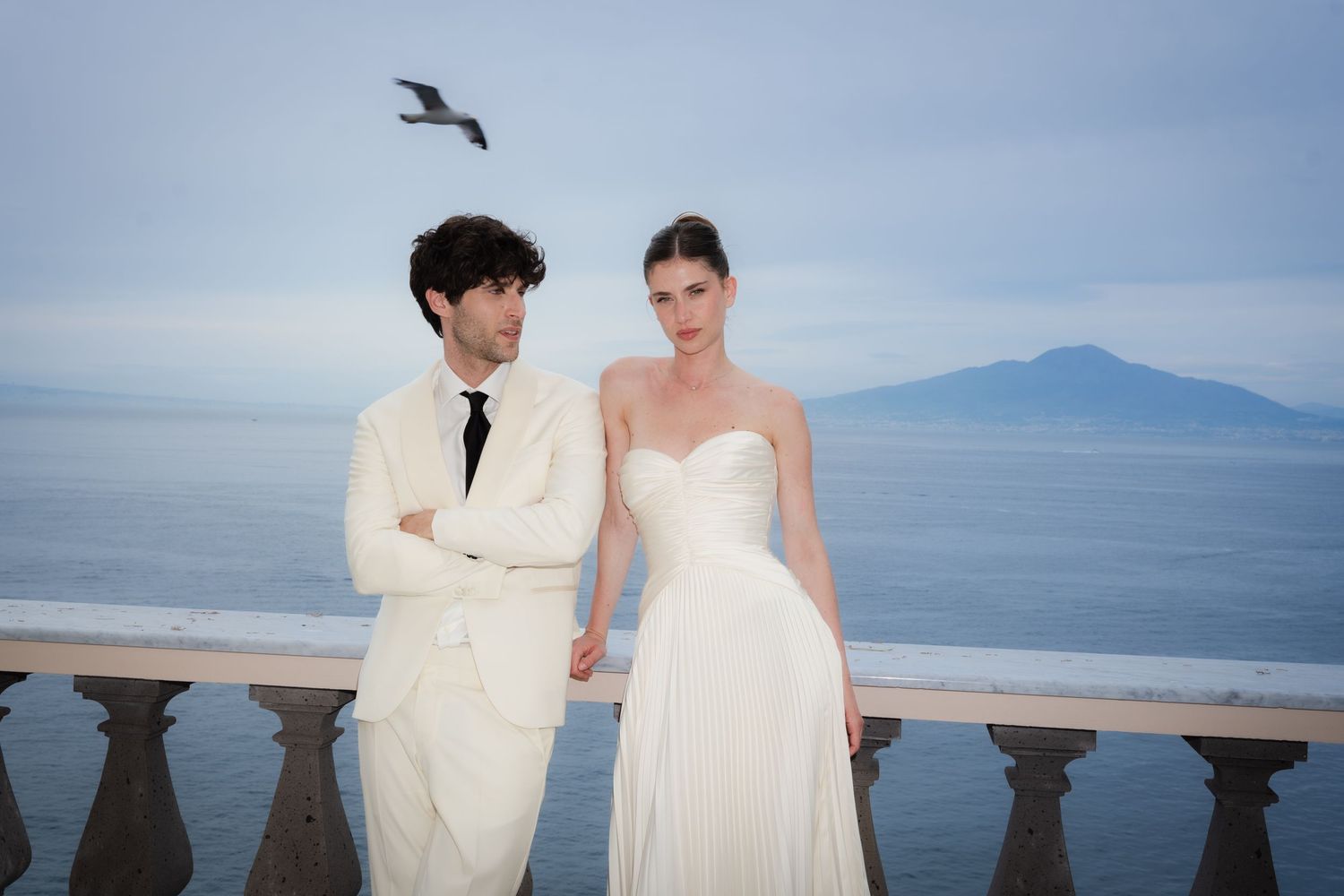 Elegant couple in white wedding attire posing on a seaside terrace with ocean and mountain views.