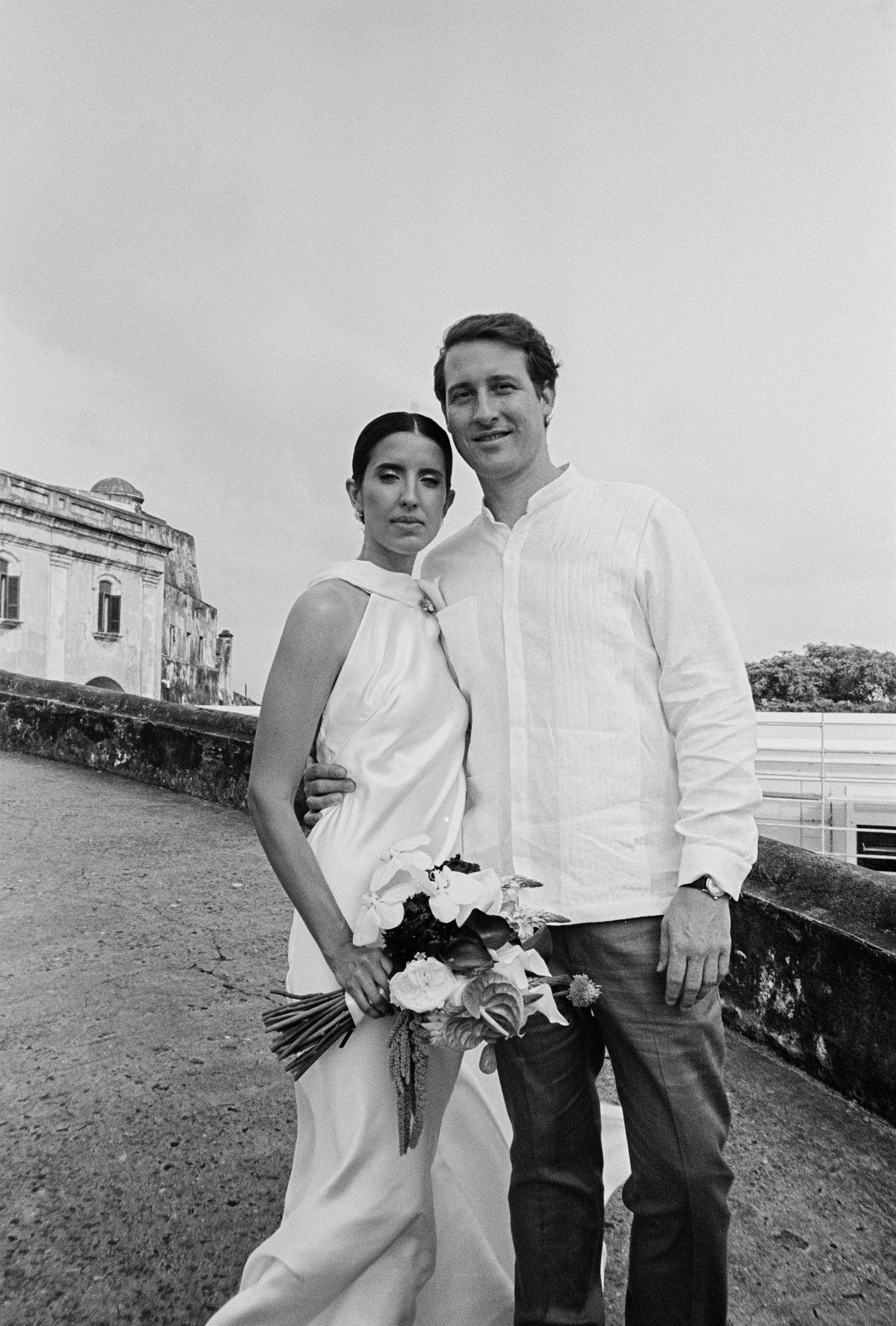 Black and white photo of a couple posing outdoors, woman in white dress holding bouquet, man in white shirt, historic building behind.