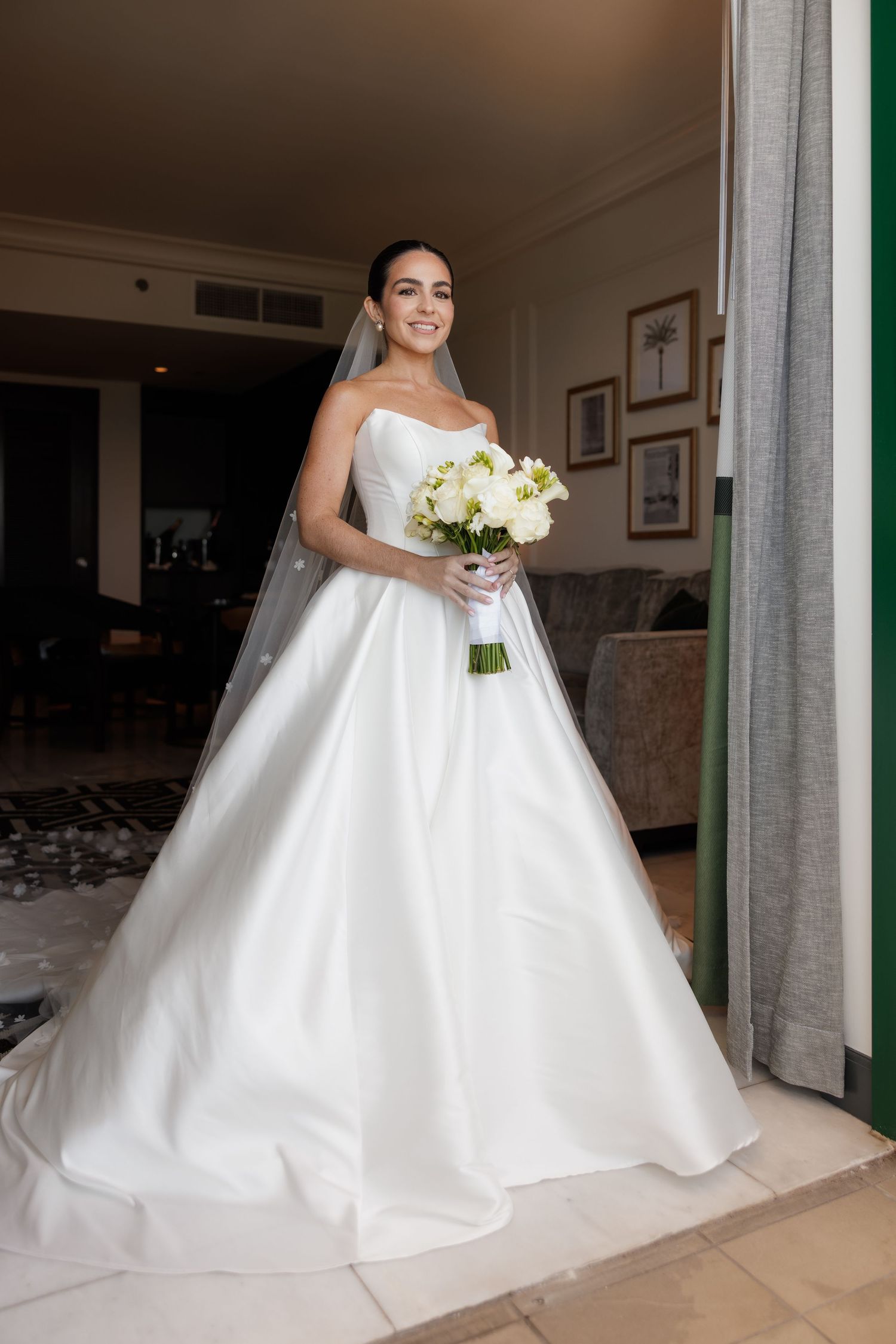 A bride in a voluminous white wedding dress stands in a room with gray curtains.
