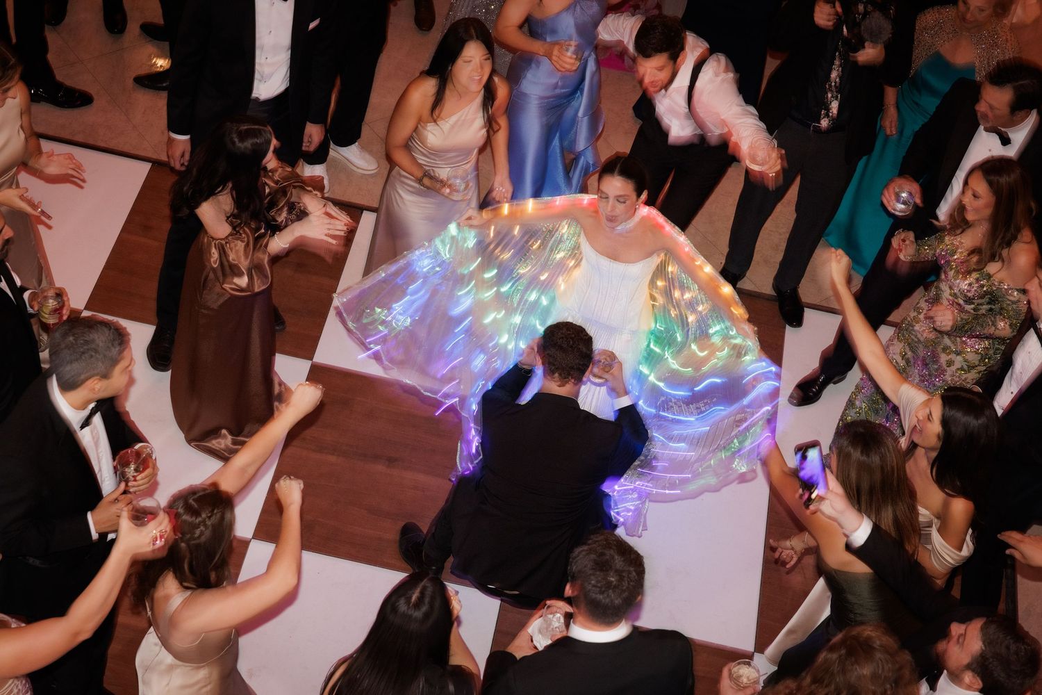 Aerial view of dancers on a ballroom floor with iridescent light trails creating a colorful glow effect.