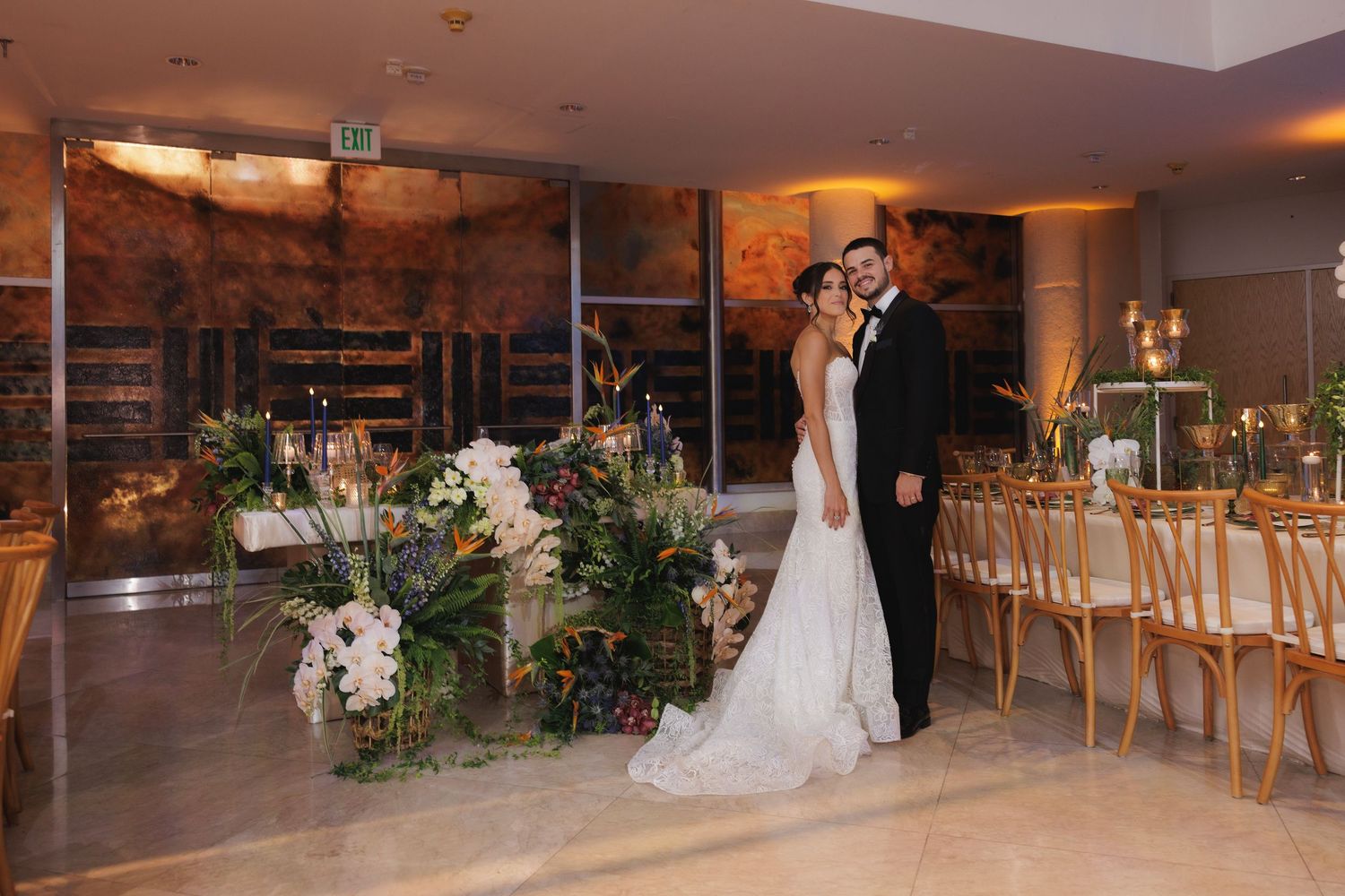 Elegant wedding reception setup in modern venue with lush greenery arrangements and gold chairs at sunset.