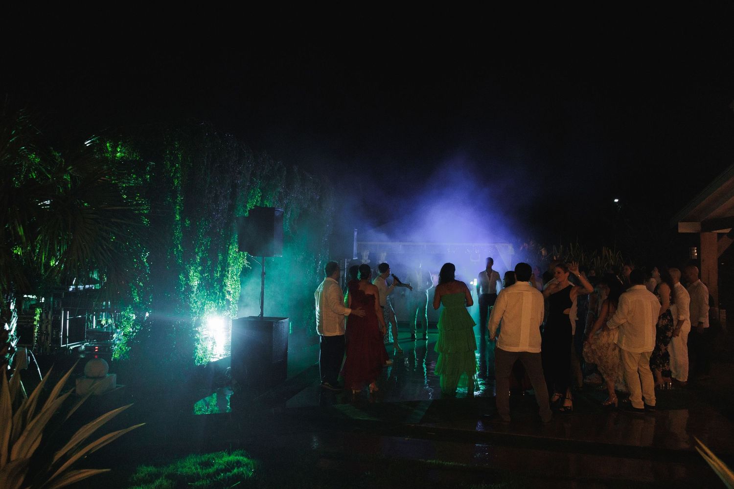 Dark outdoor scene with green lighting illuminating trees and people walking in the distance.