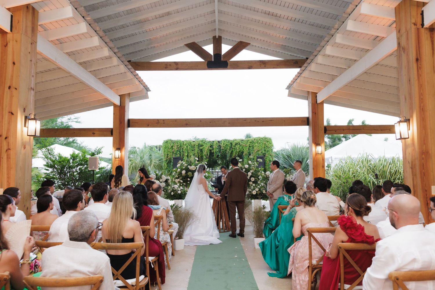 Outdoor wedding ceremony under a wooden pavilion overlooking lush green landscape with guests seated on white chairs.