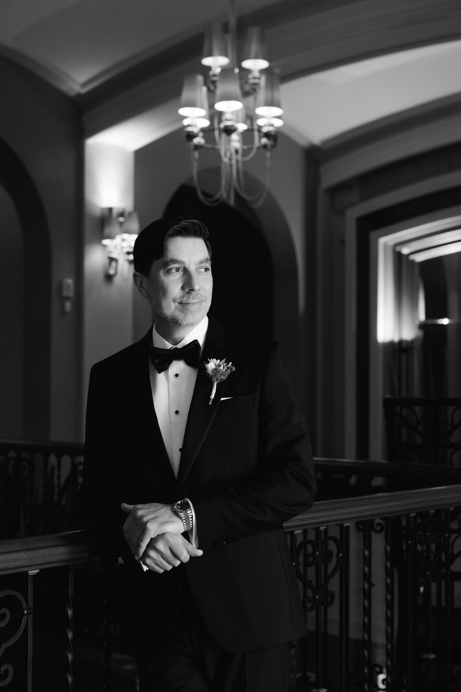 Black and white portrait of person in formal tuxedo standing in elegant hallway with chandelier lighting.