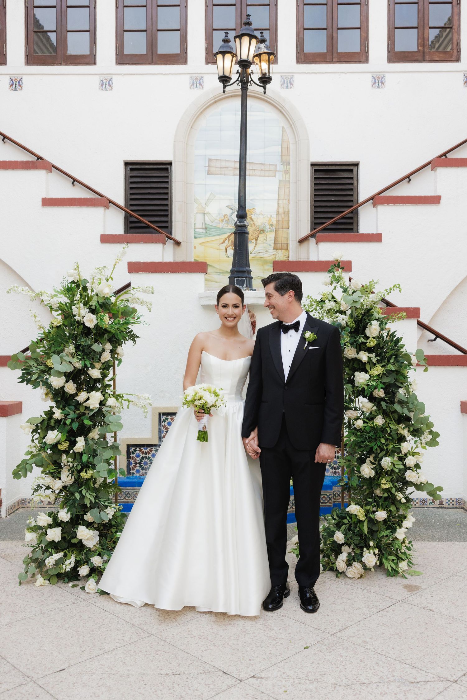 A wedding couple poses under an elegant archway decorated with white floral arrangements and a vintage lamp.