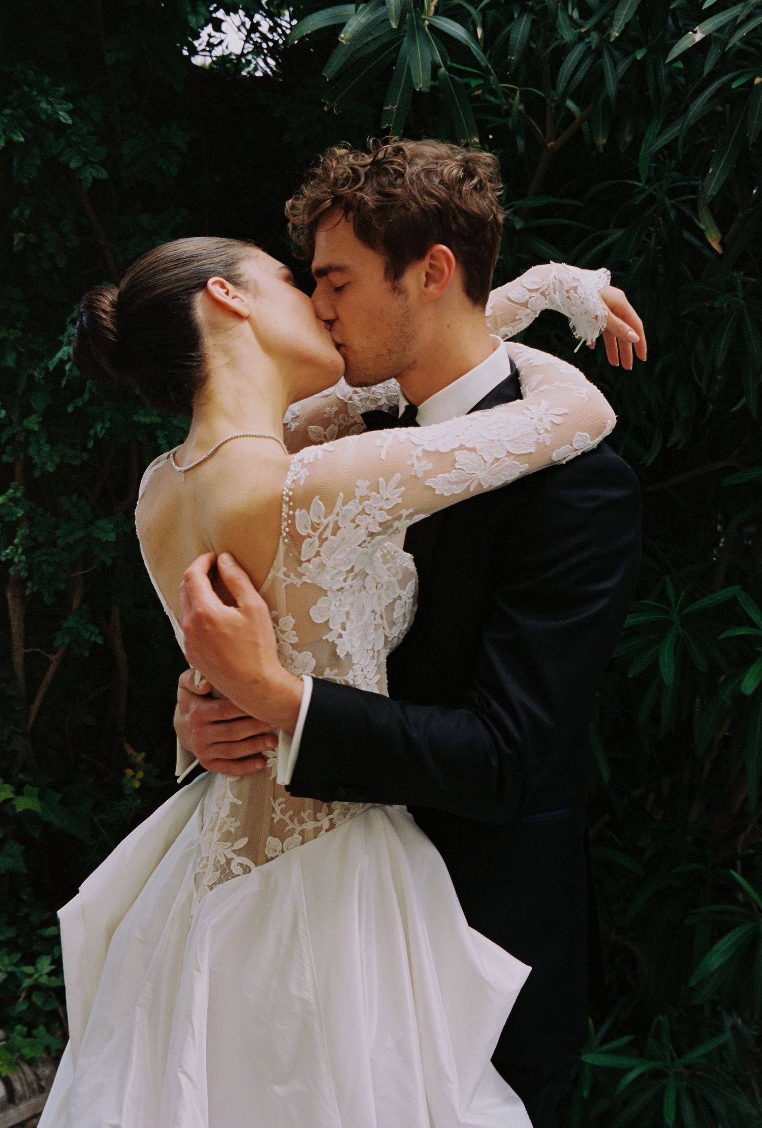 A bride and groom share a romantic kiss in a lush garden, she wearing a lace wedding gown.