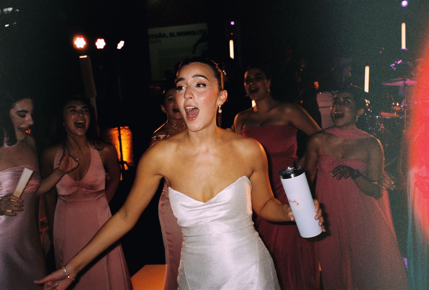 Bride in white dress dancing energetically with bridesmaids in pink at a wedding reception.