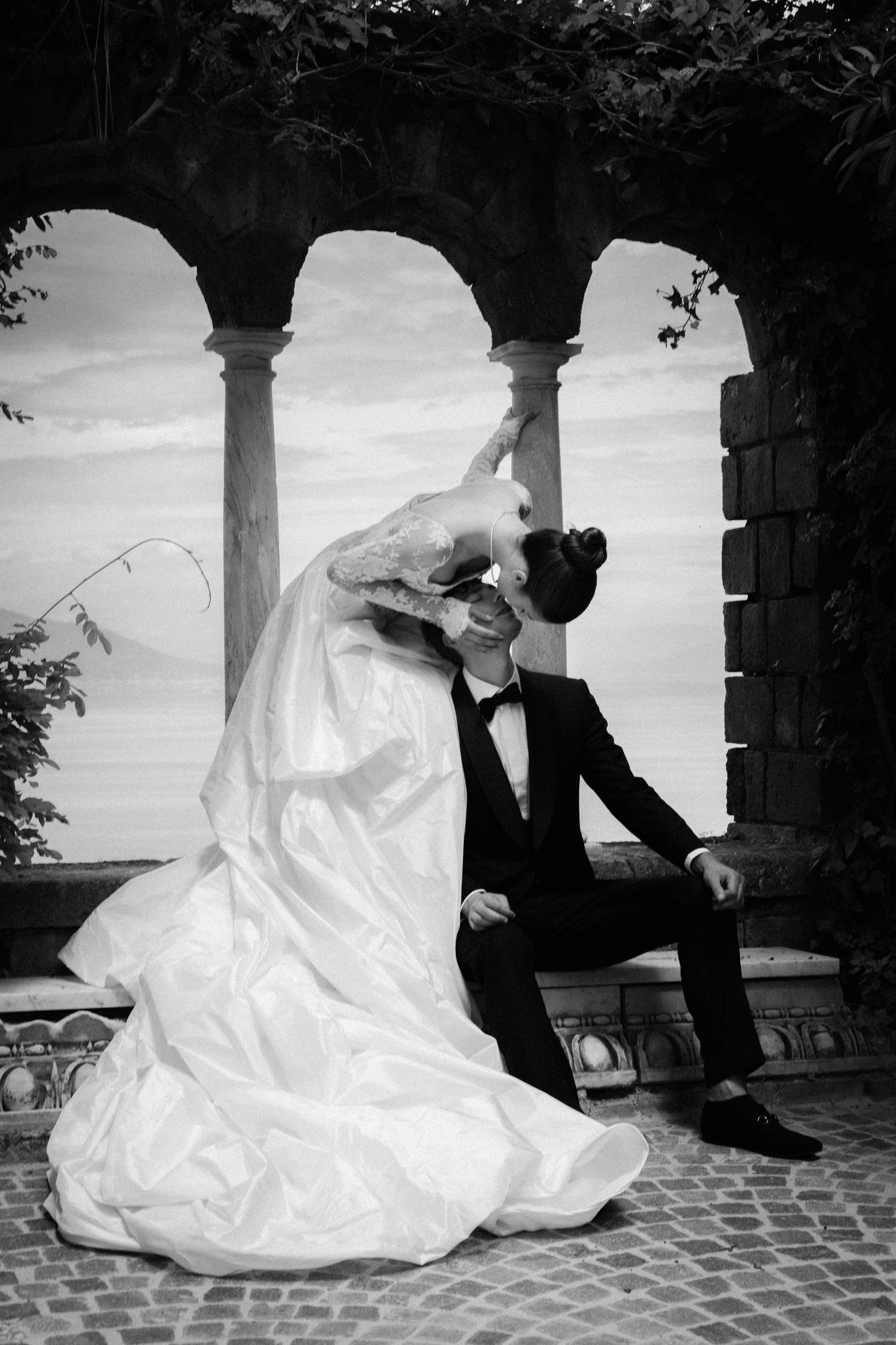 Black and white photo of a bride and groom sharing a romantic kiss under stone arches near a lake.