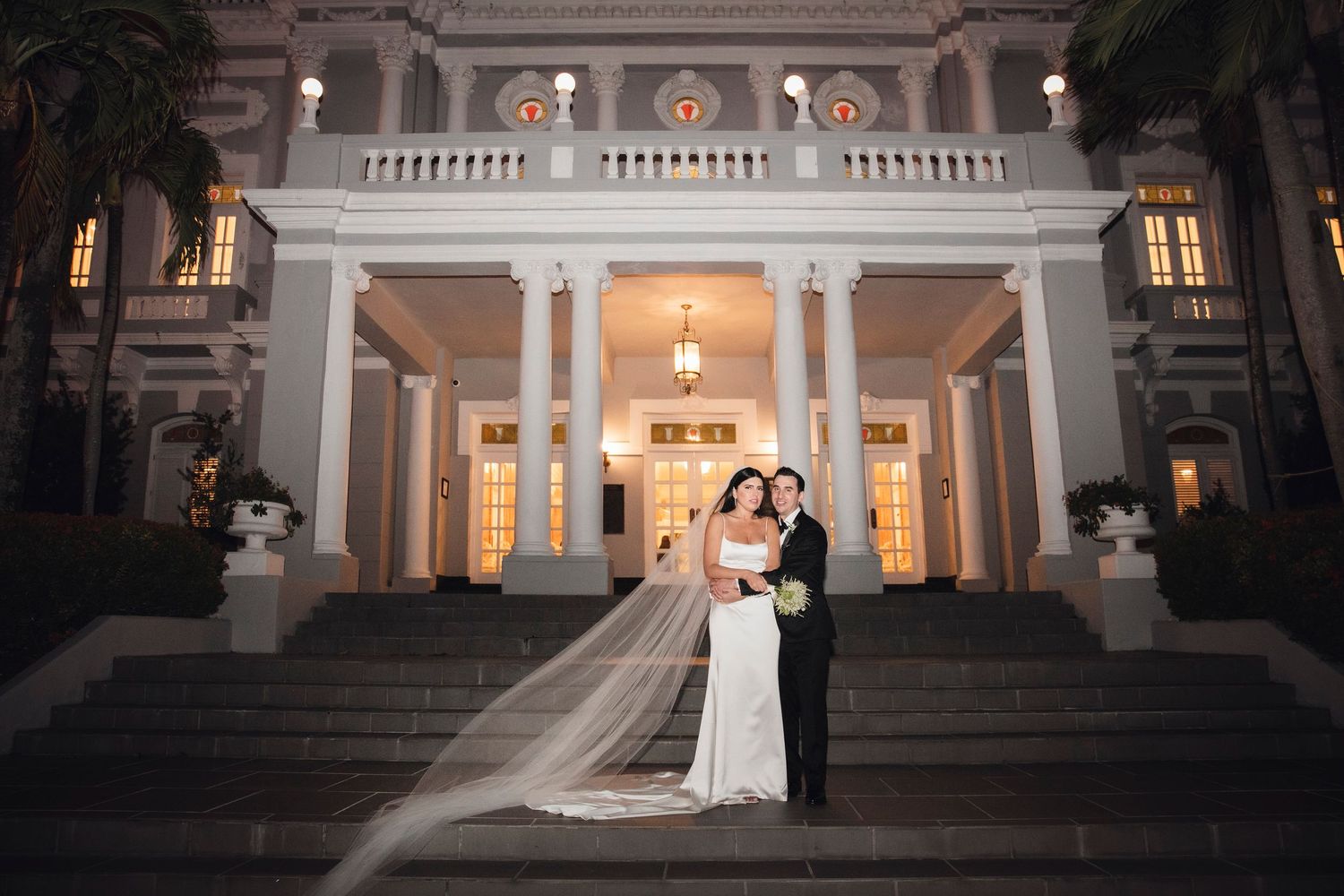 Elegant couple poses at dusk in front of a grand white columned mansion with glowing interior lights.