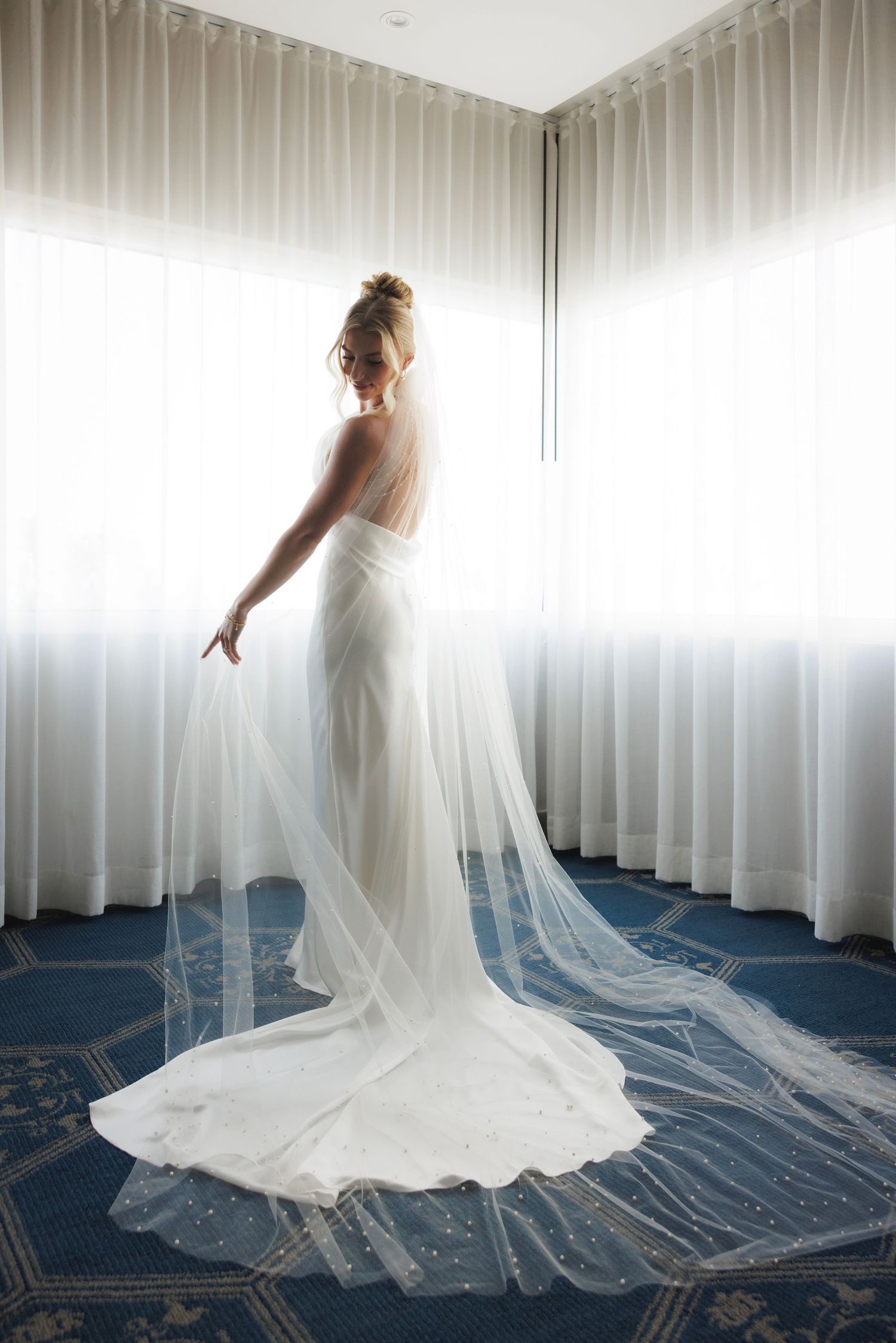 A bride in a flowing white veil gown poses gracefully against a backdrop of white curtains in a wedding venue.