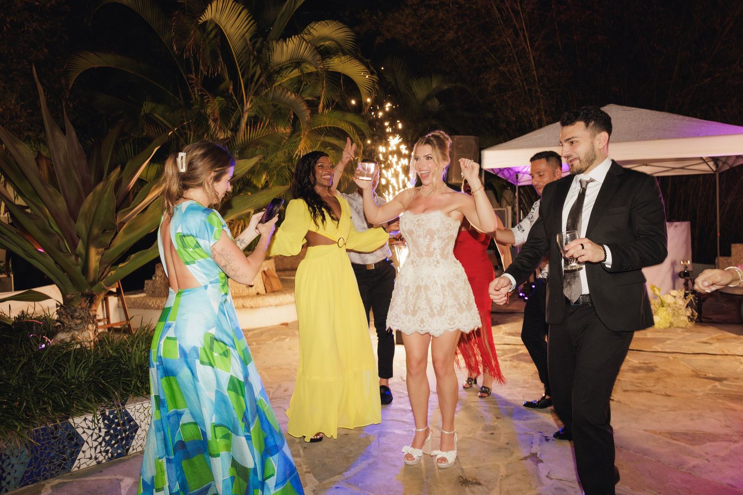 People in formal attire dancing and socializing at an outdoor evening party with tropical plants in the background.