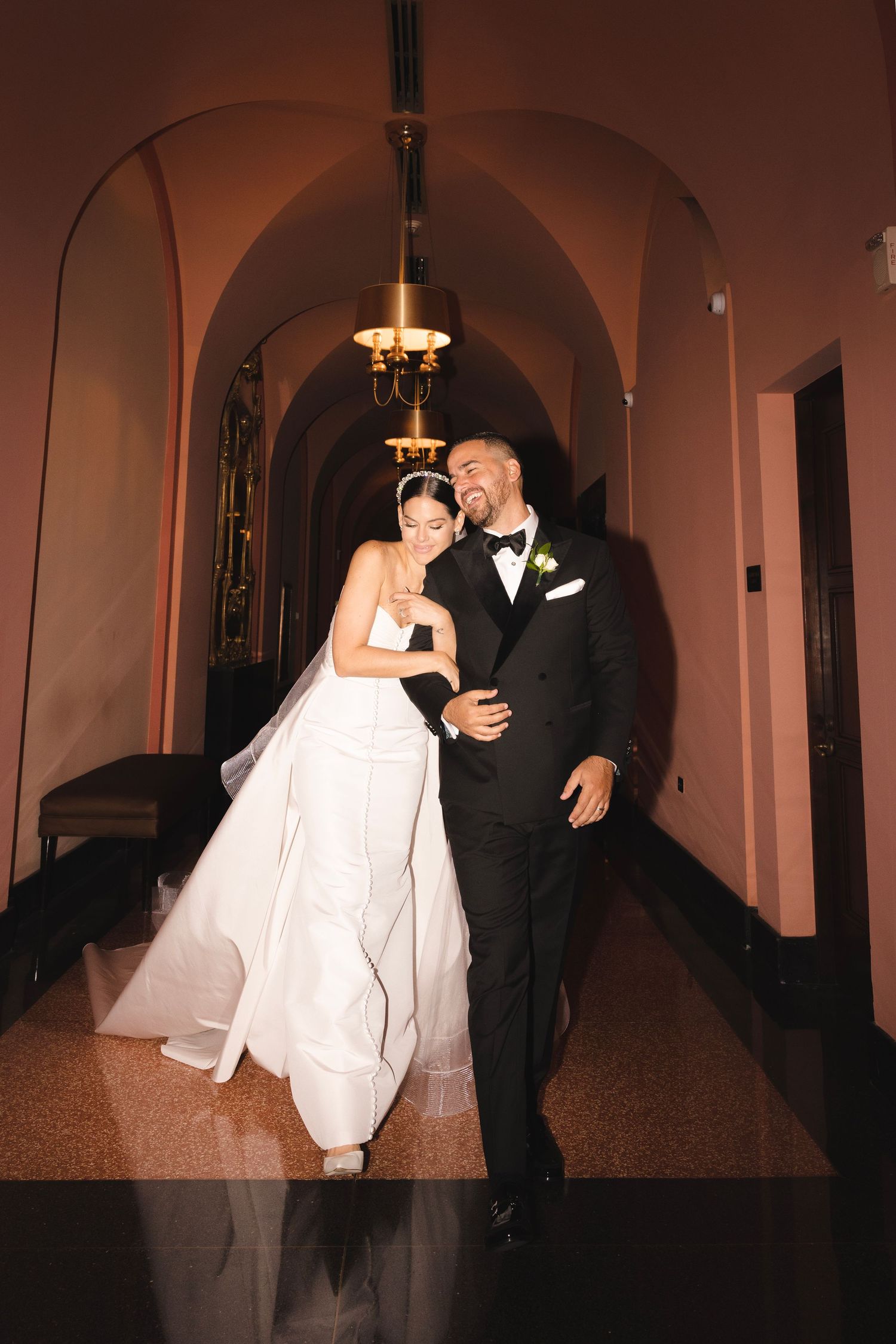 A bride and groom share a joyful moment together while walking down an elegant hallway with arched ceilings.