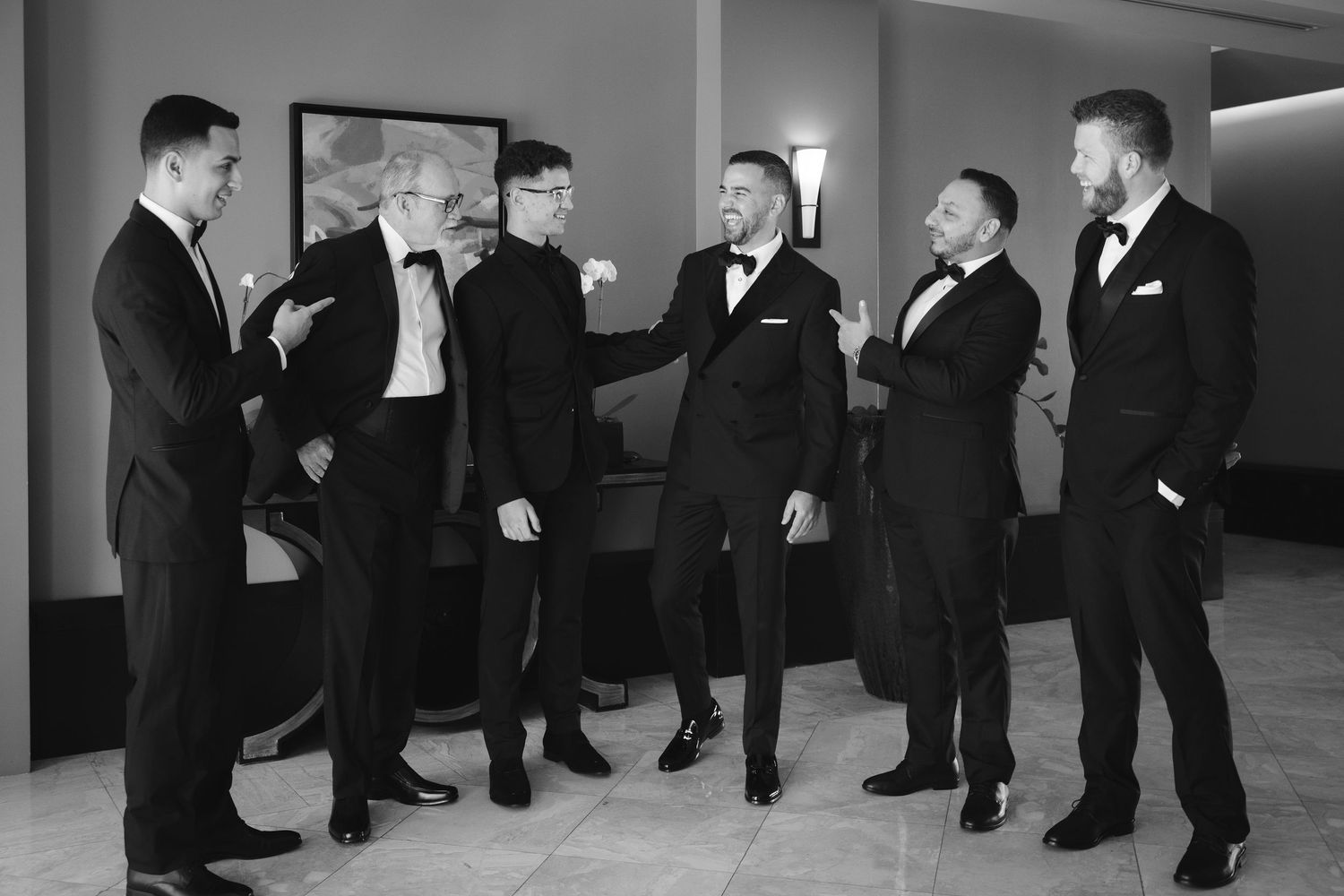 A group of groomsmen in black tuxedos line up against a gray wall for a formal wedding photo shoot.
