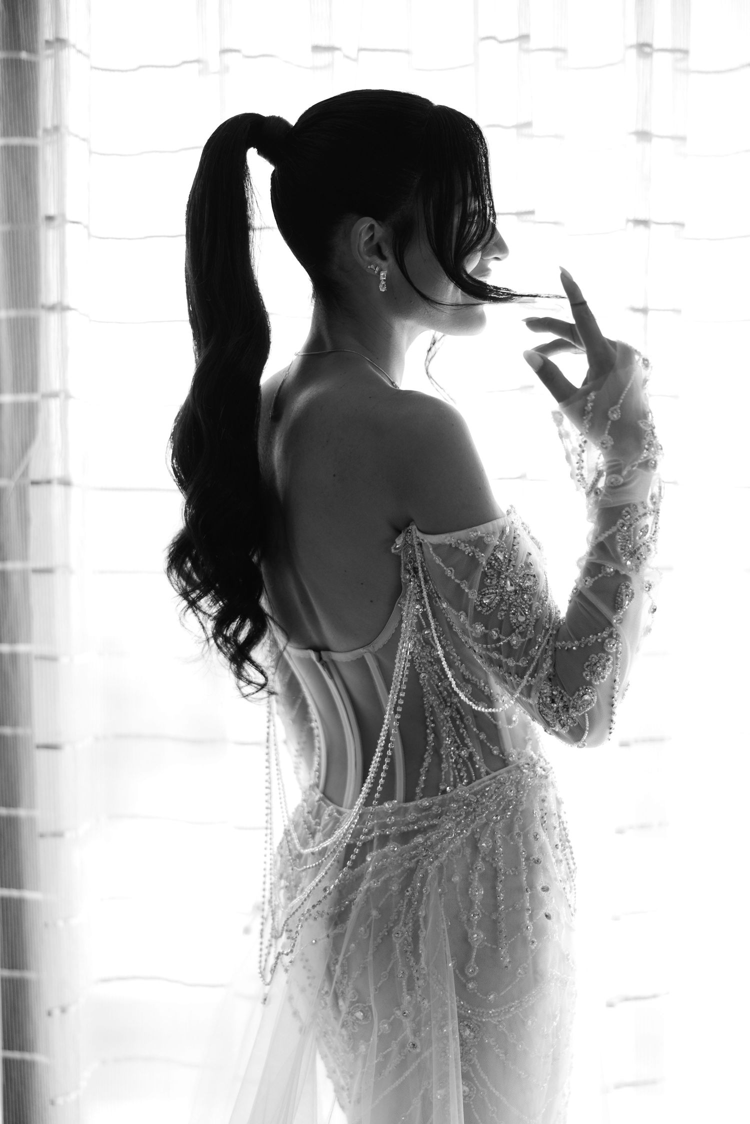 Silhouette of a bride in a beaded wedding gown with long ponytail against window light.