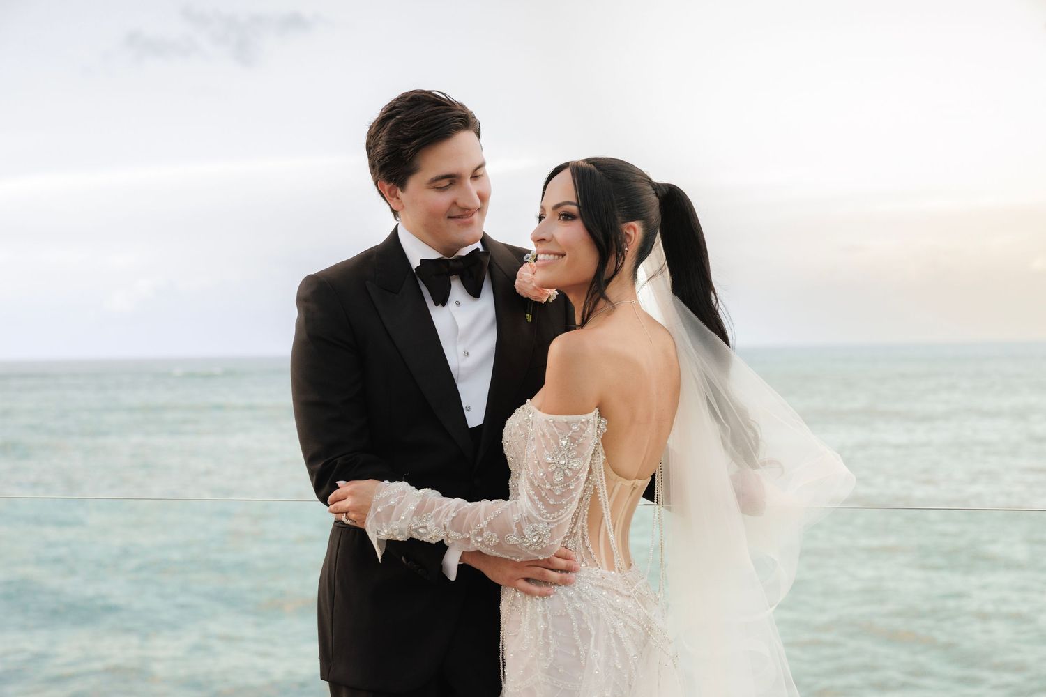 Newlywed couple embraces on beach with turquoise ocean in background during seaside wedding ceremony.
