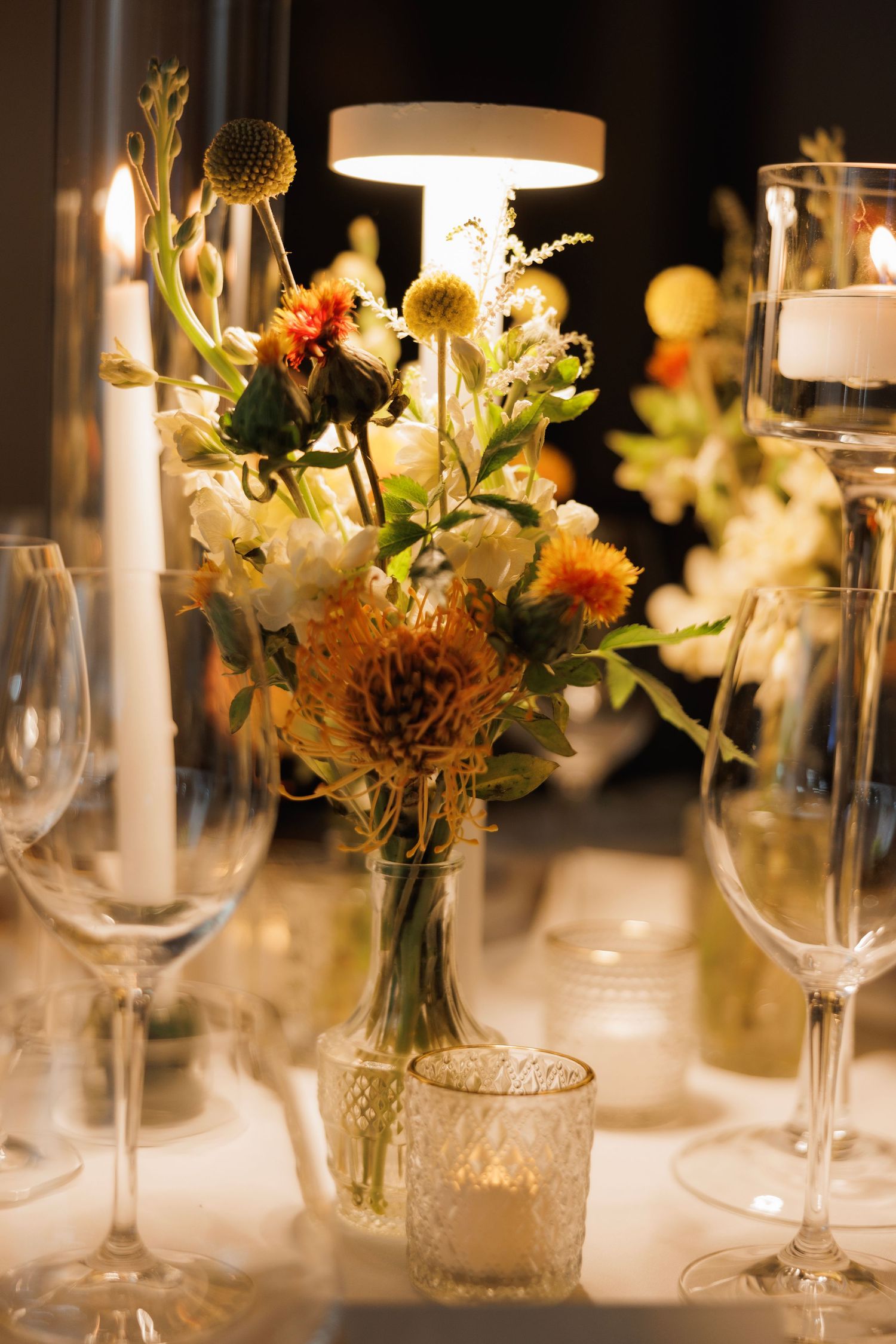 Wedding reception table setting with elegant wine glasses and delicate floral centerpieces in glass vases at night.