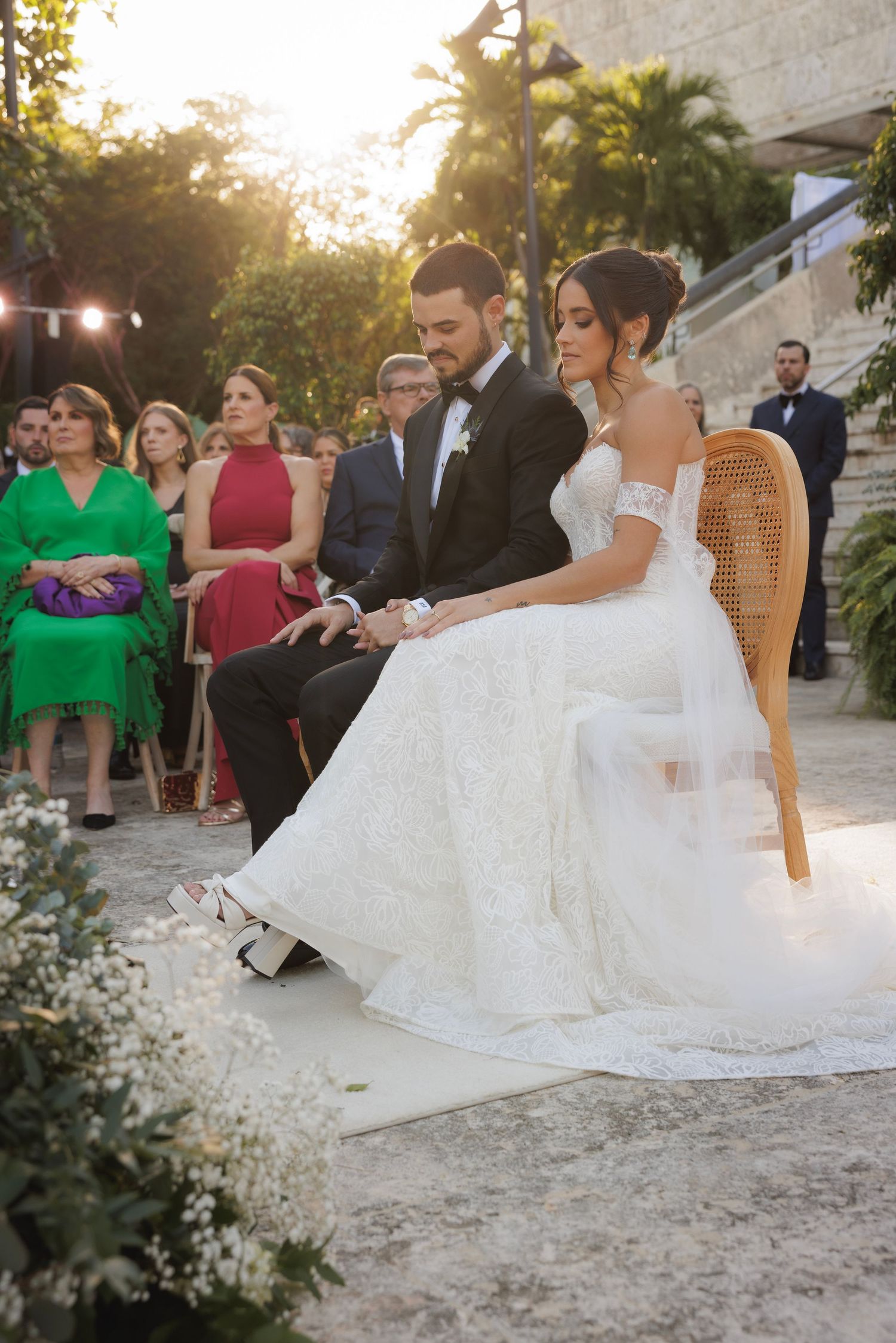 A romantic outdoor wedding scene with rattan chairs and guests during golden hour sunset.