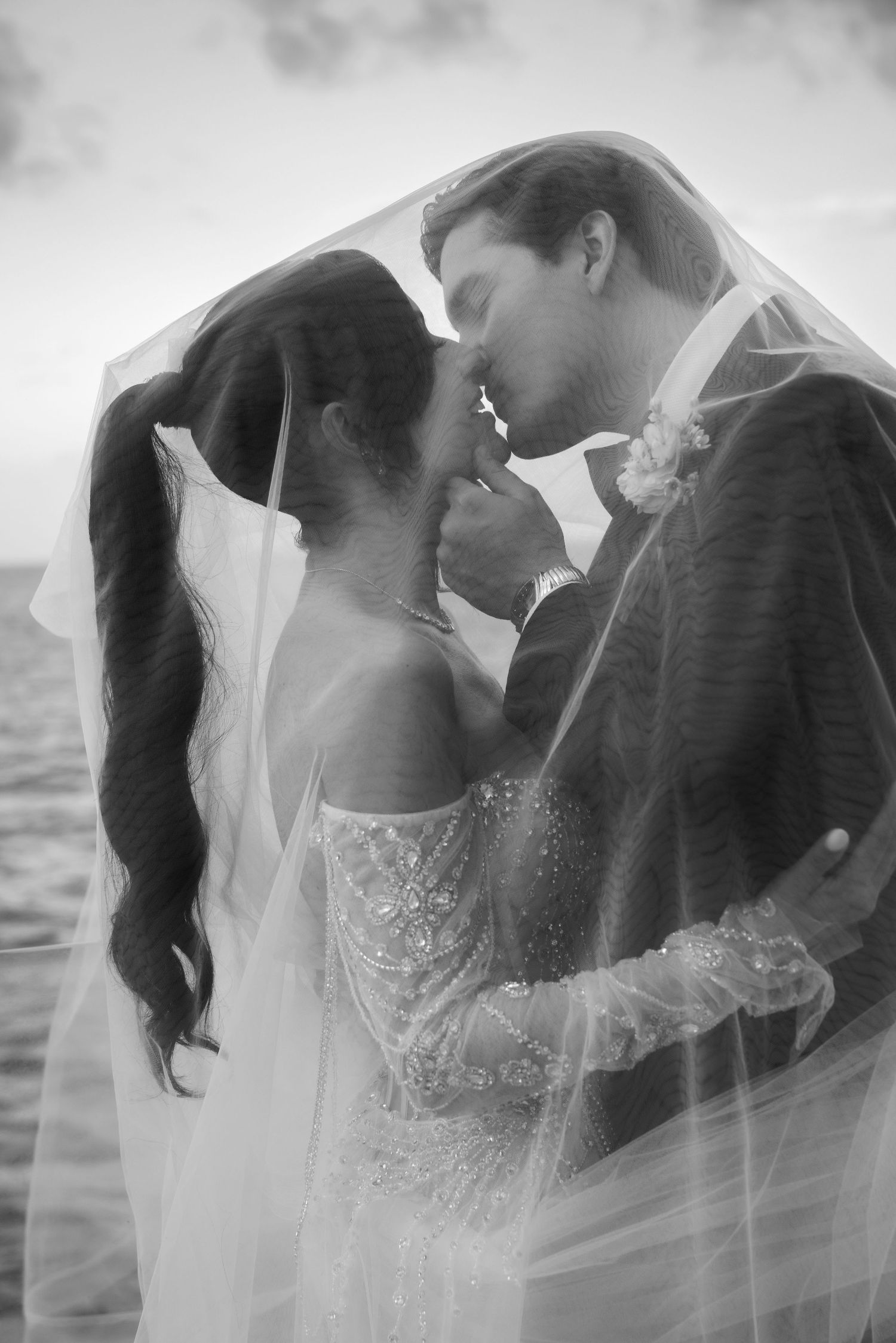 A black and white romantic wedding photo series showing a couple embracing under a flowing veil at sunset.