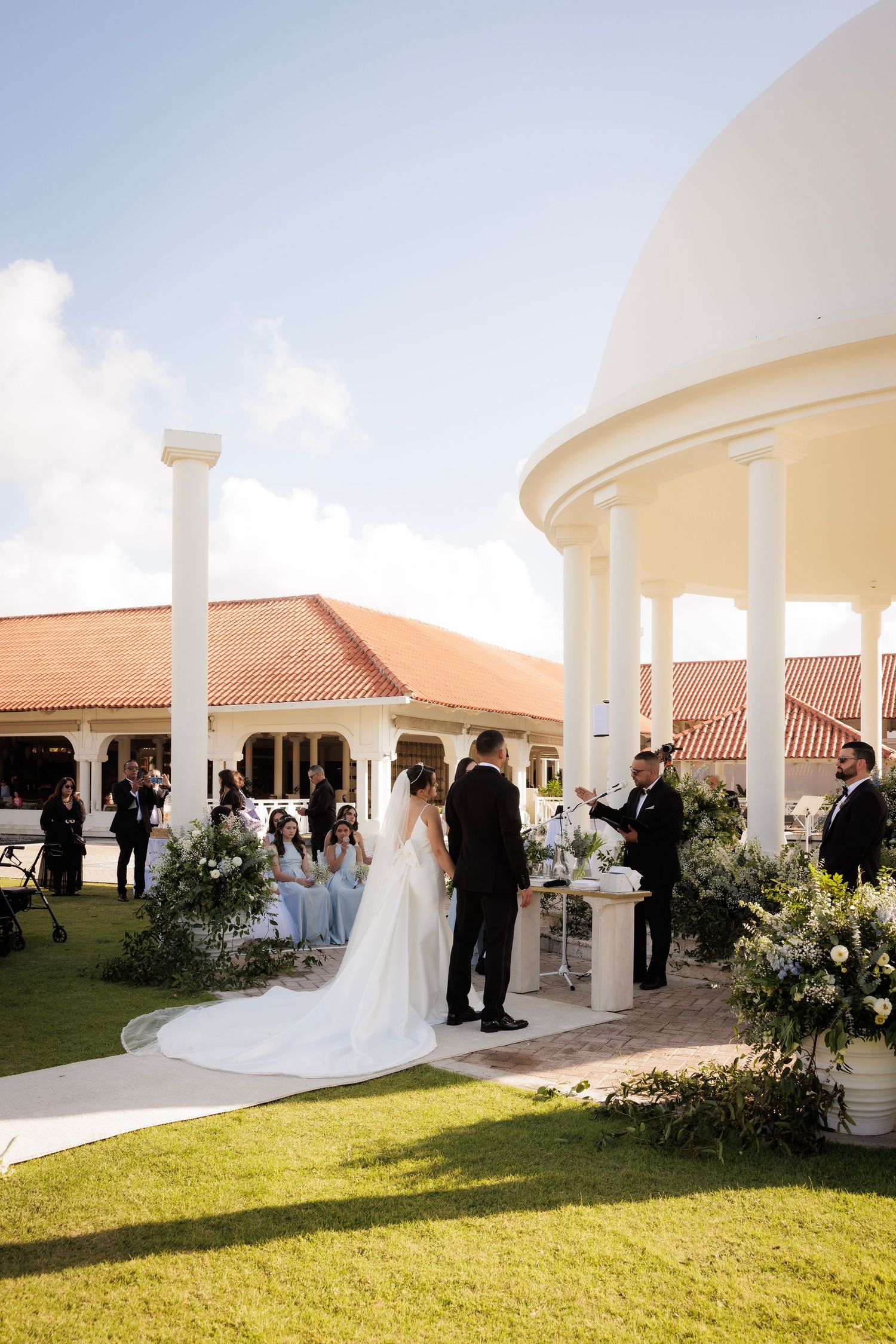 Wedding ceremony taking place at an elegant white gazebo with Mediterranean architectural details near a sunny lawn.