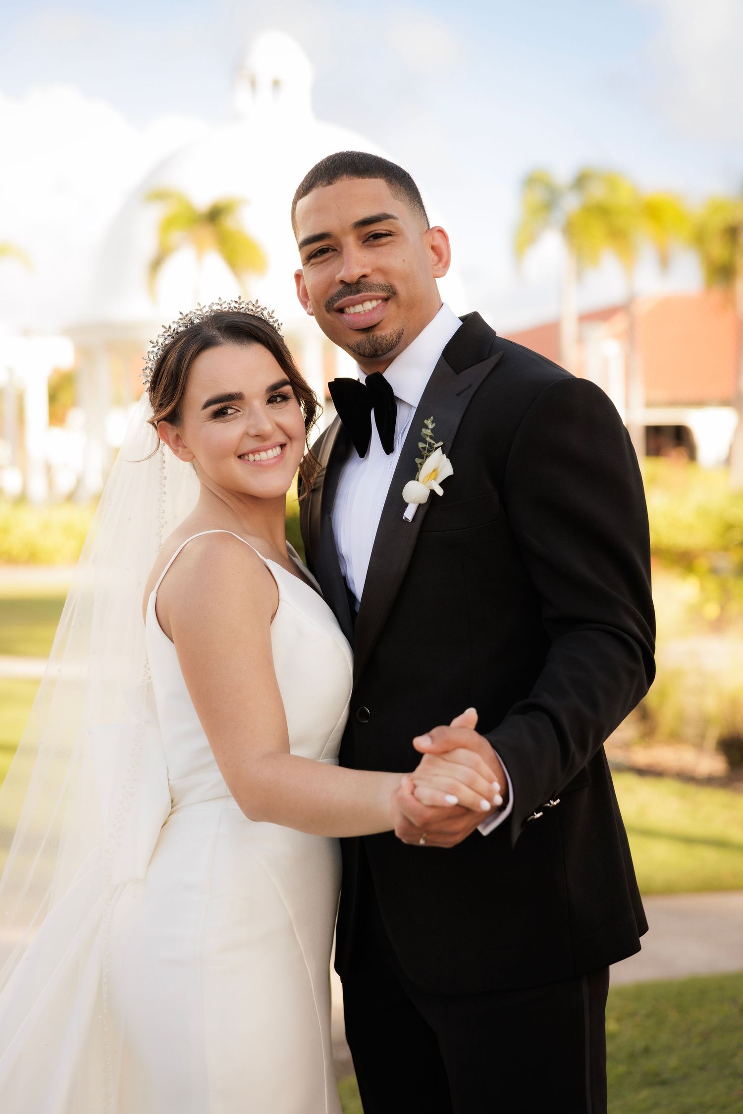A happy bridal couple shares romantic dance poses together on their wedding day against a sunny palm tree backdrop.