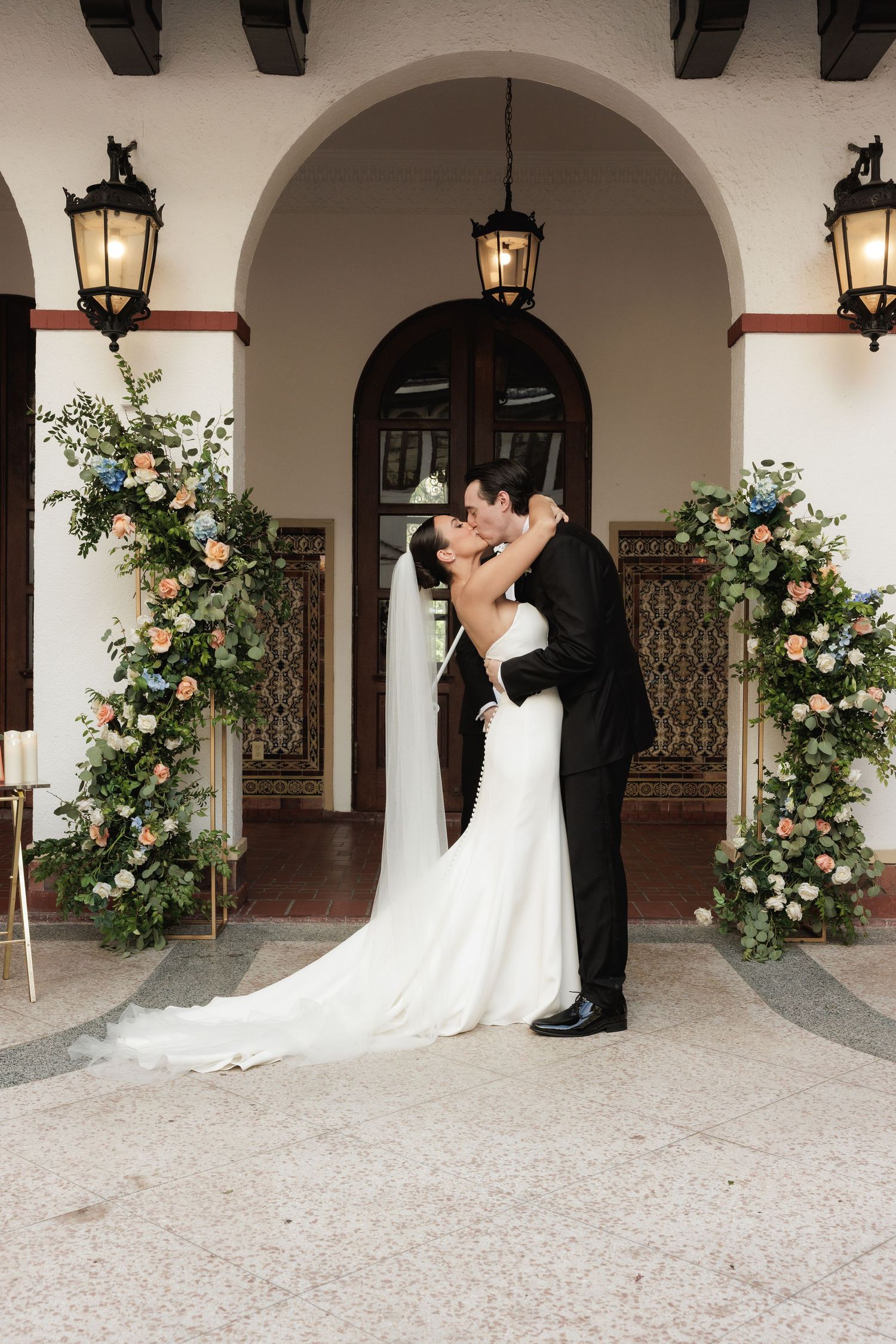 Romantic kiss in Spanish colonial archway decorated with elegant floral arrangements and vintage lanterns.
