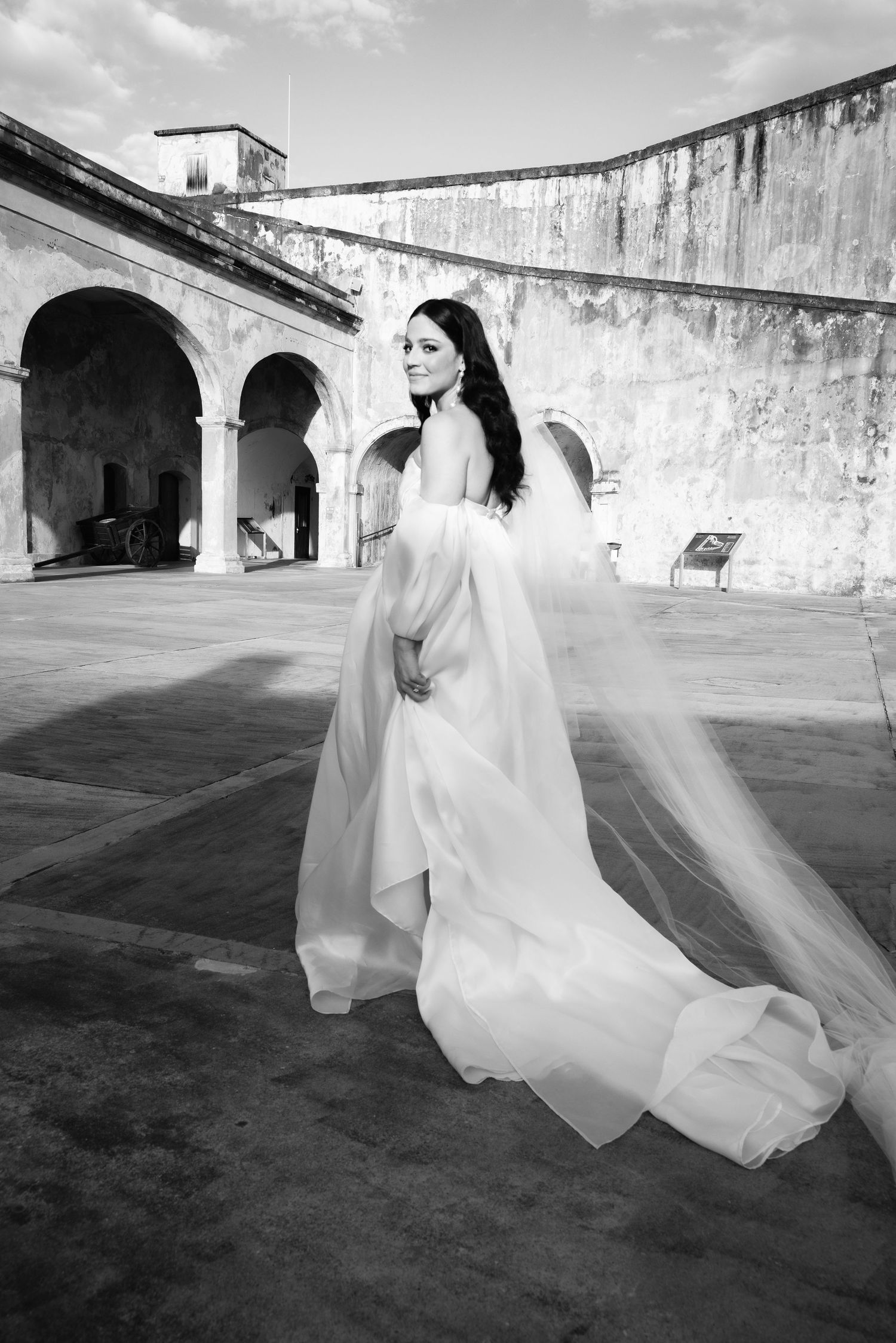 Wedding dress flowing dramatically against historic stone architecture with arched colonnade.