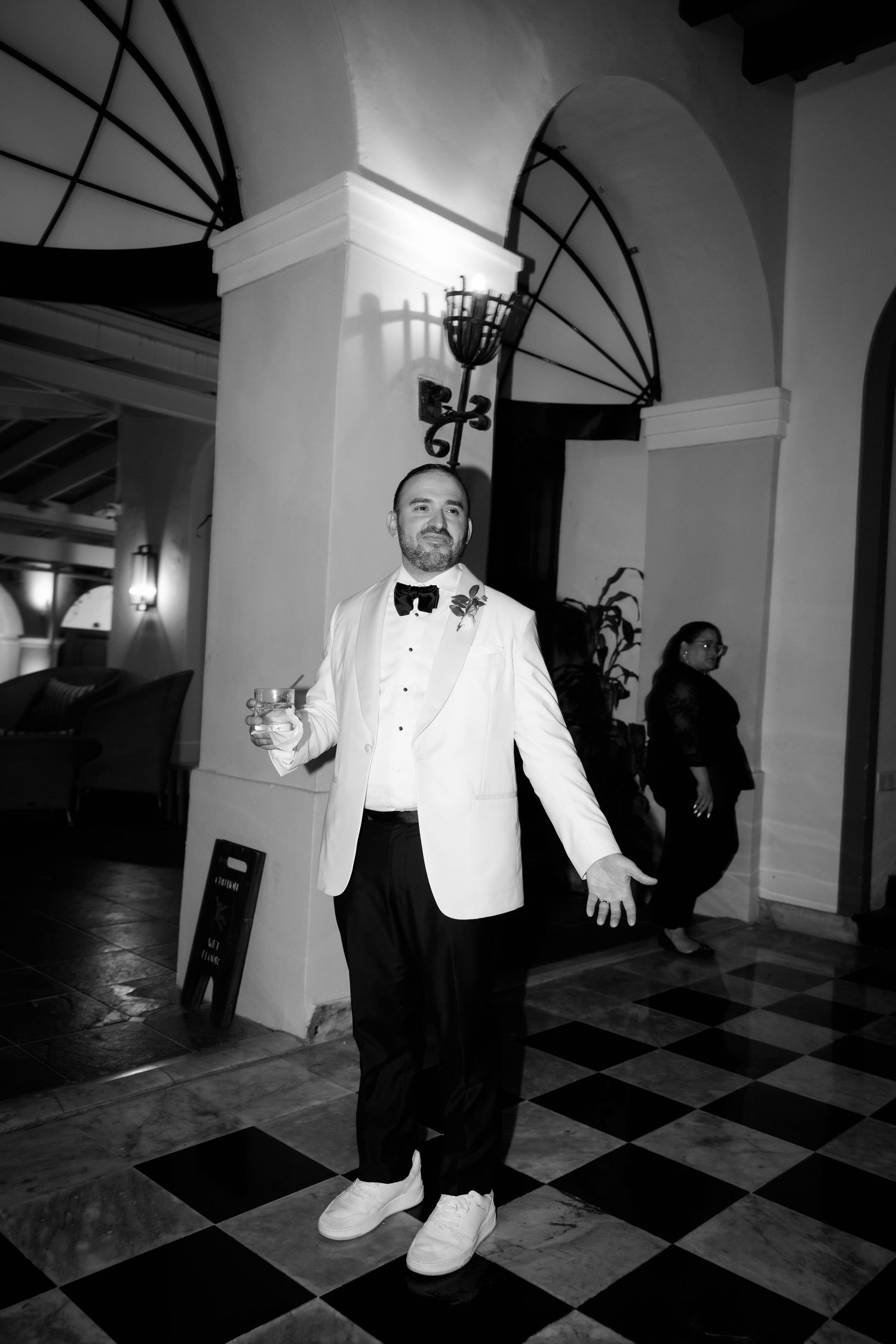 A person in a white tuxedo jacket and black pants dances on a black and white checkered floor in a grand hallway.