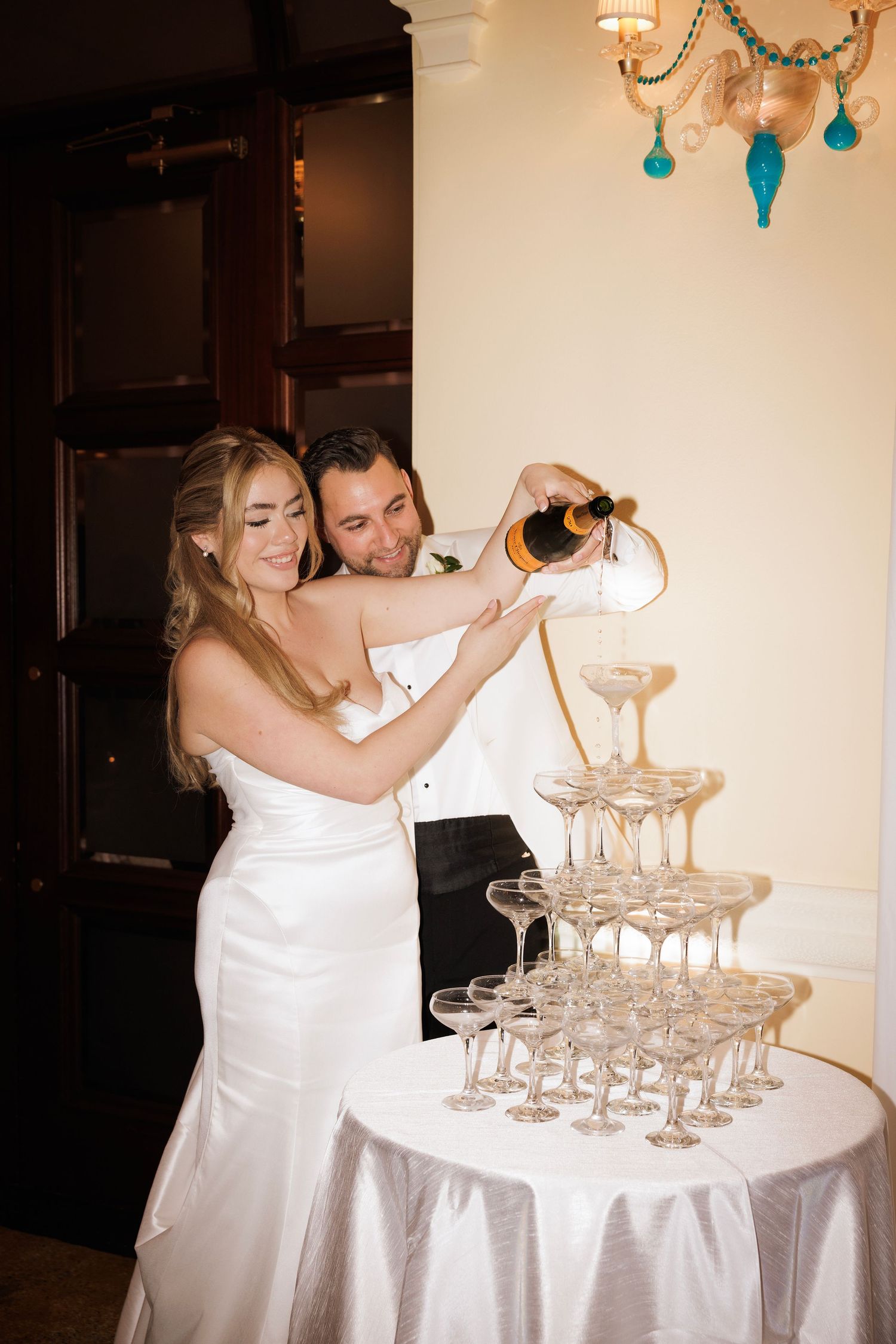 A couple pours champagne into a tower of elegant glass coupes at a formal event.