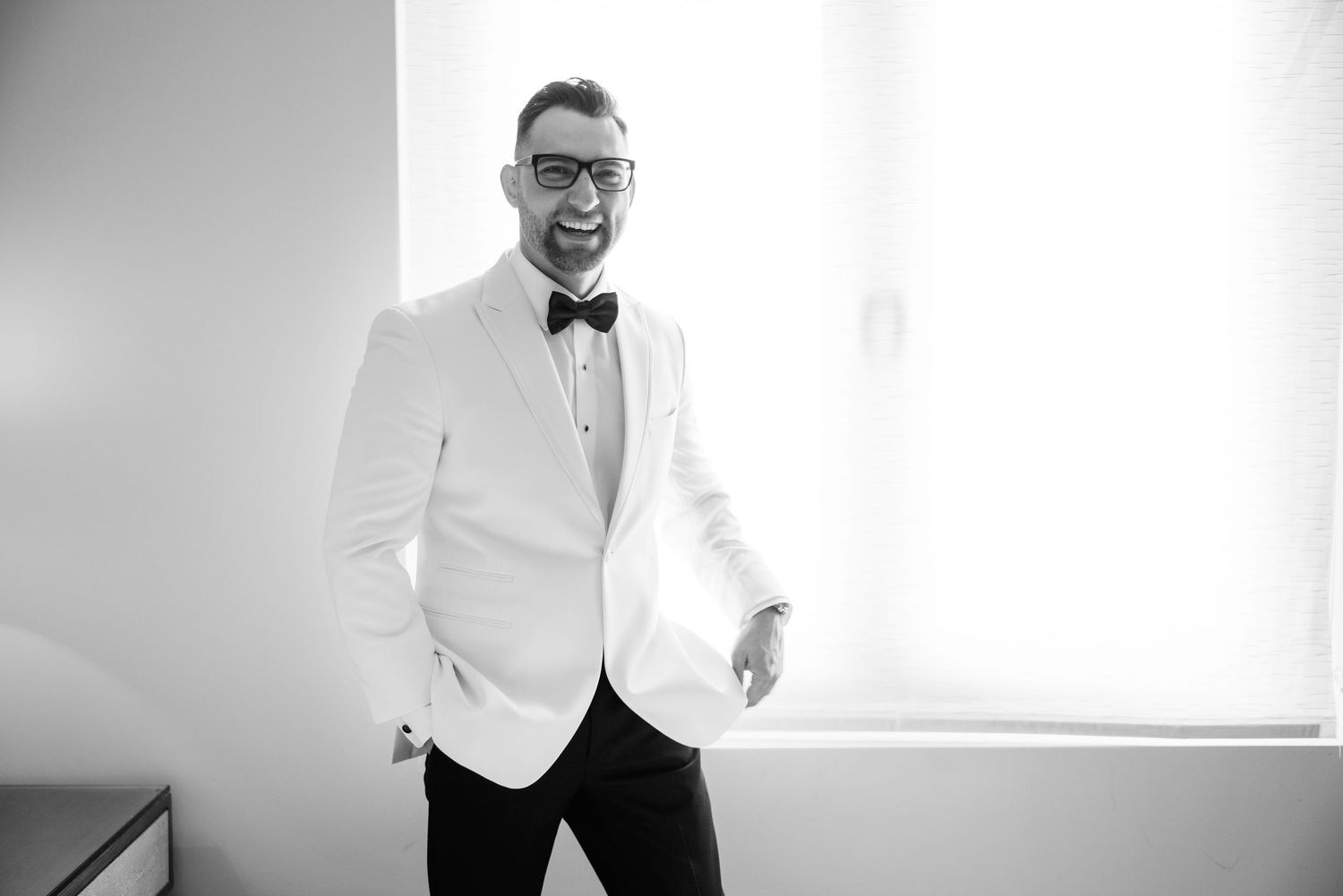 A groom in a white tuxedo jacket and bow tie poses in a bright minimalist room.