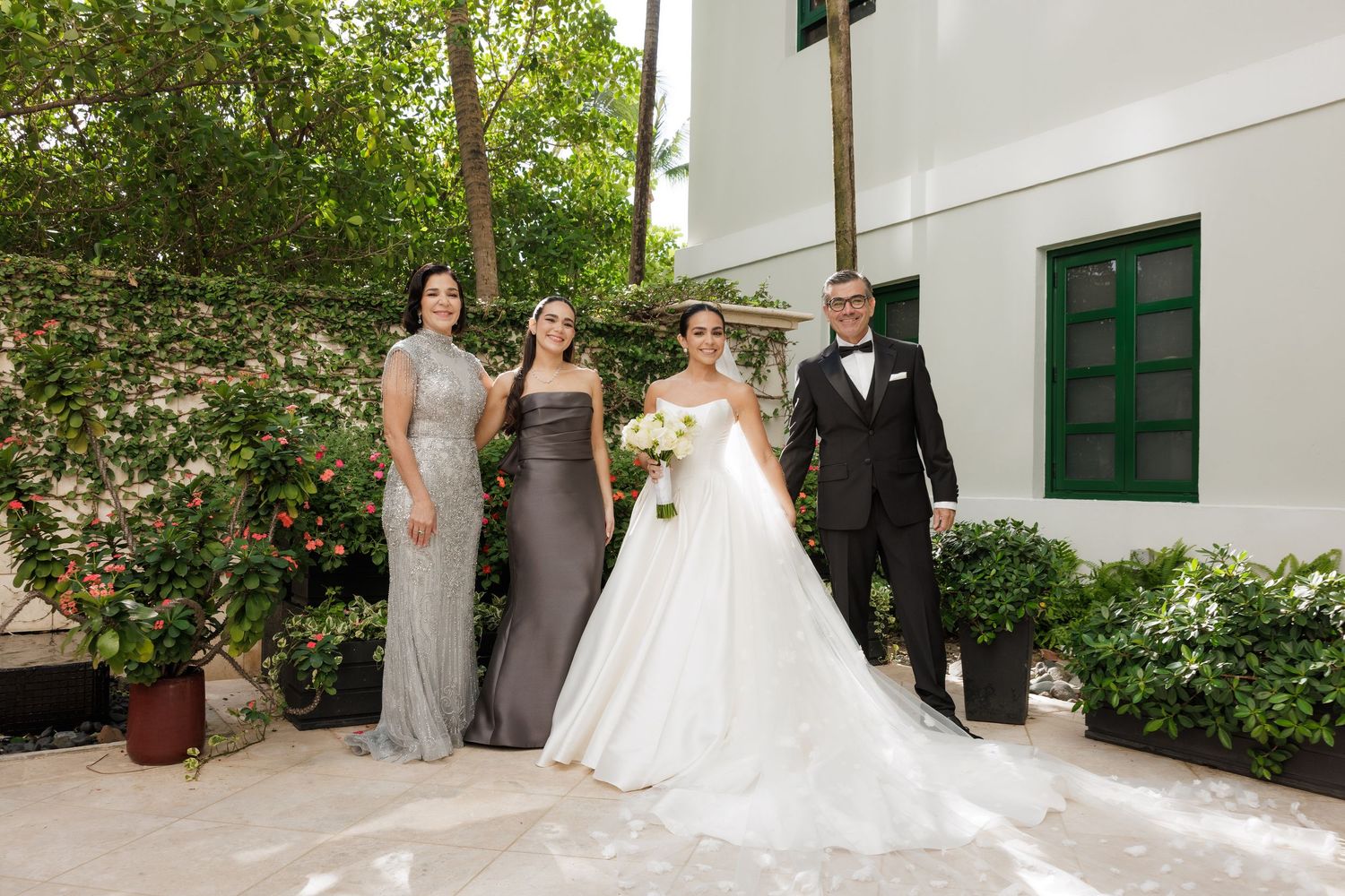 A wedding party poses together in a garden setting with elegant formal attire and flowing white gowns.