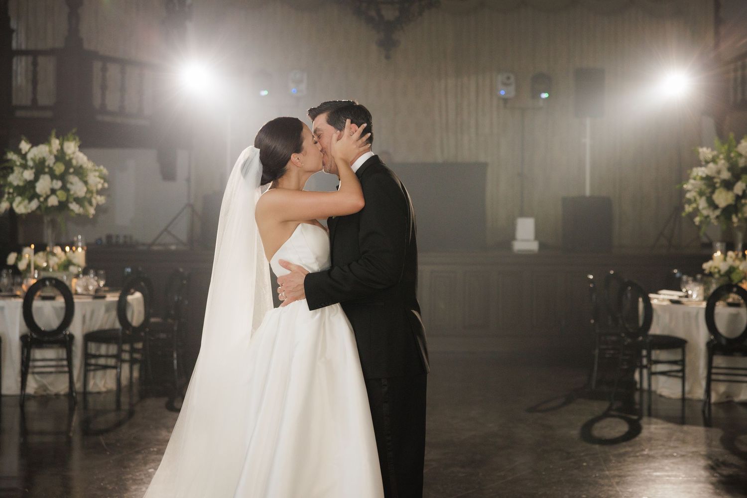 Romantic couple embraces on a dimly lit dance floor with elegant wedding decor and soft lighting in the background.