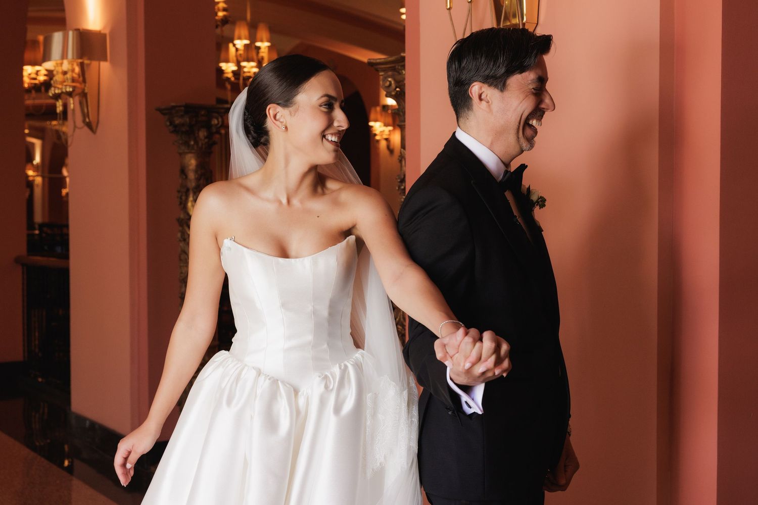 A bride in a white strapless gown and groom in black tuxedo share a first look moment in a hallway.