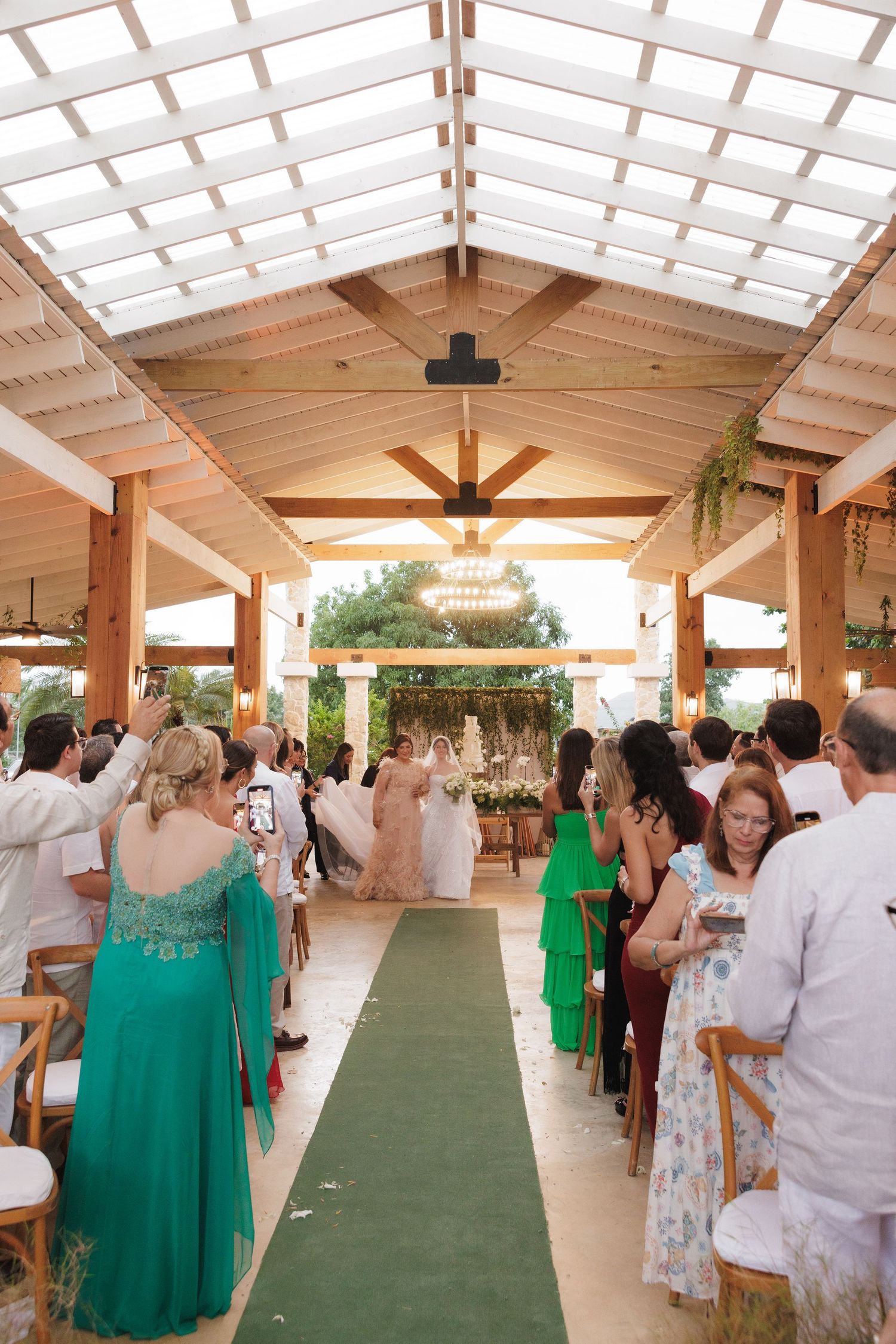 Wedding guests gather in an elegant covered pavilion with a green aisle runner and natural lighting from skylights above.