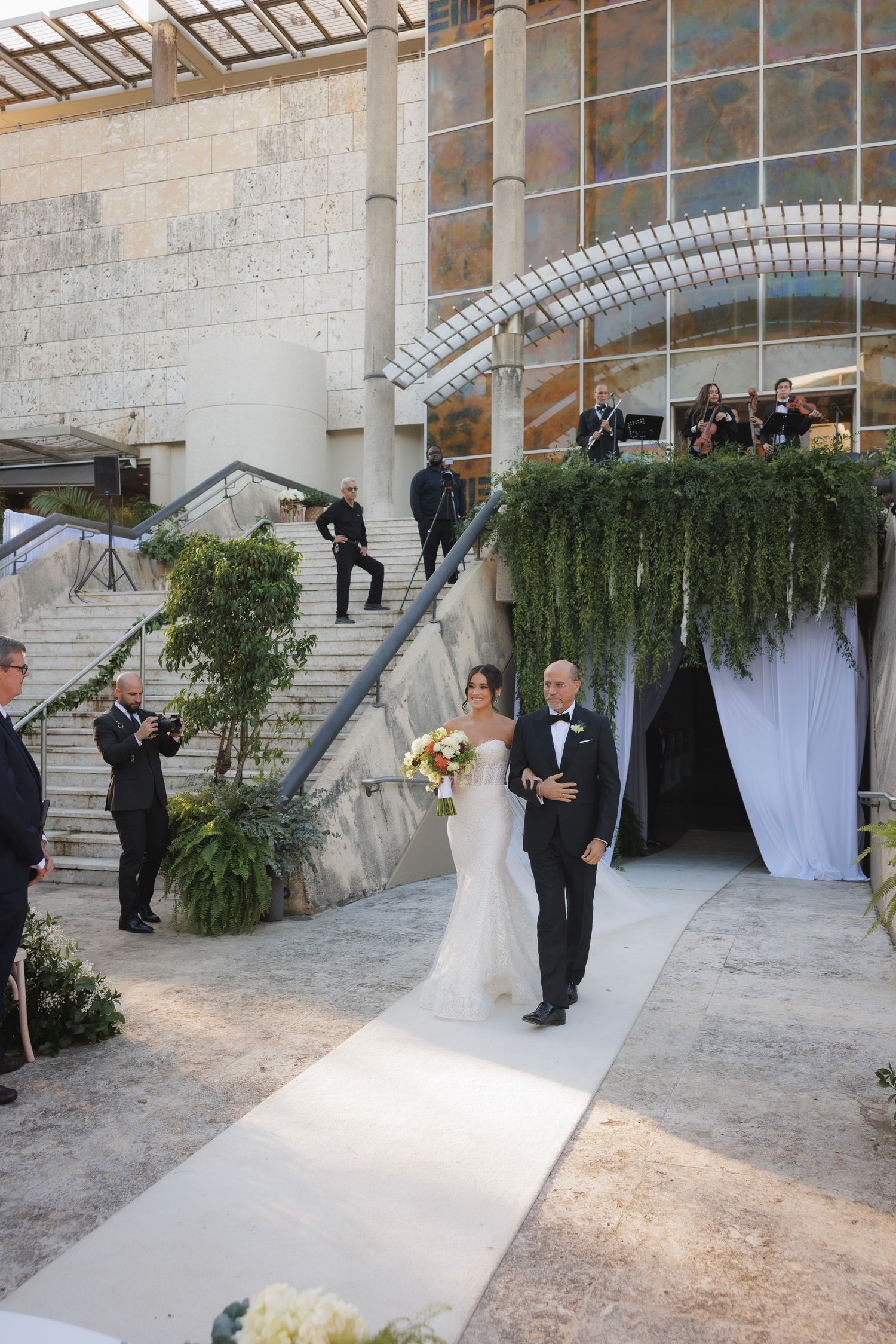 Outdoor wedding ceremony setup with white draping and greenery against stone architecture.