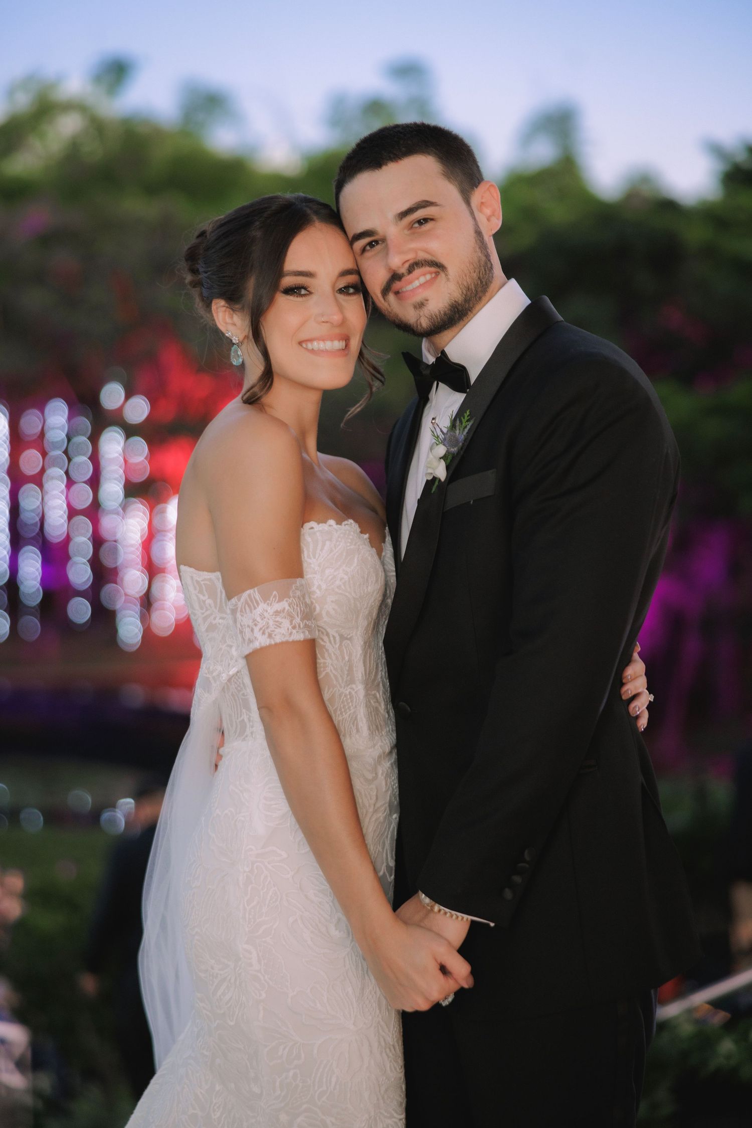 A romantic wedding couple poses together outdoors at dusk with soft bokeh lights and floral decor in the background.