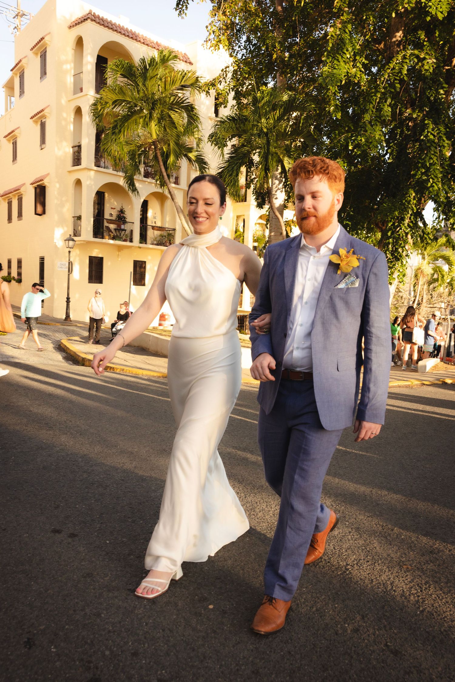 A couple walks together on a palm tree-lined path outside a Mediterranean-style building.